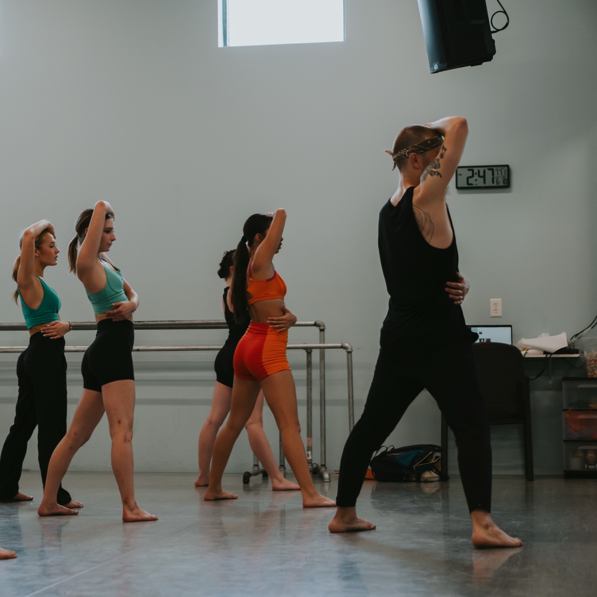 Group of five women standing in a dance studio, some with hands on their heads, one woman in front with hand on her head, all barefoot, studio with a window and a clock showing 2:41.