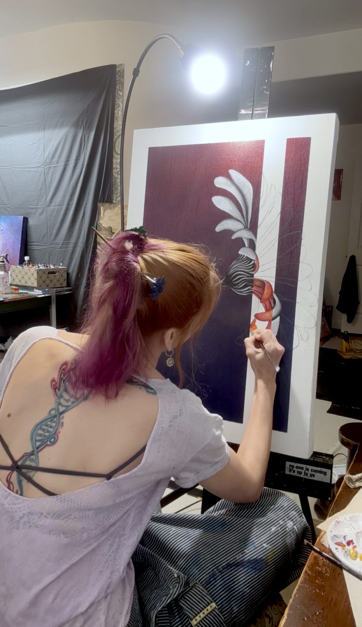 Person with purple and orange hair painting on a large canvas in a room, with a computer and other items nearby.