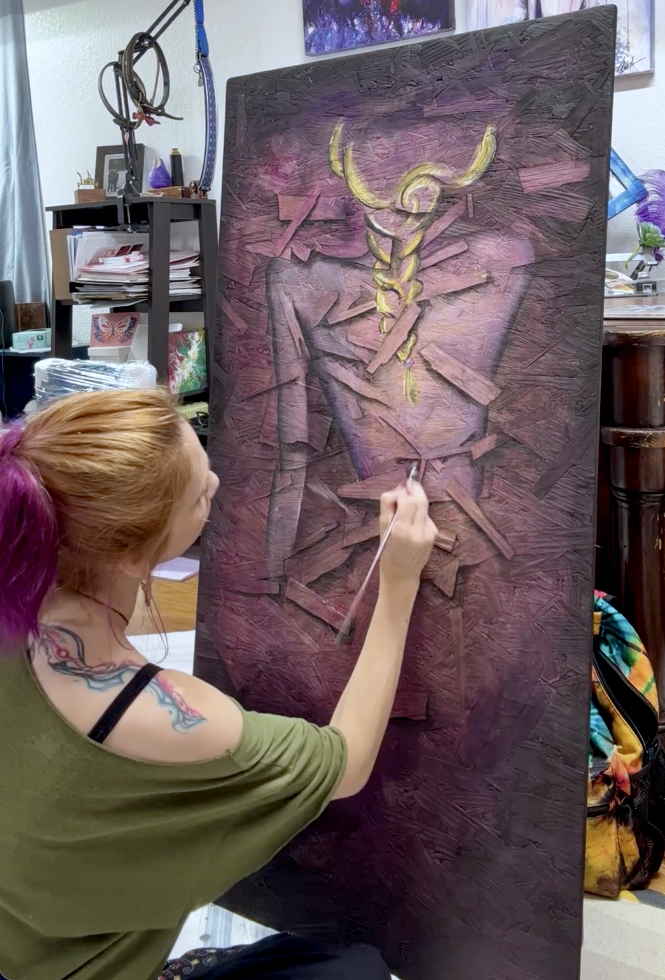 Artist with pink hair painting a large abstract portrait on a vertical canvas in a studio.