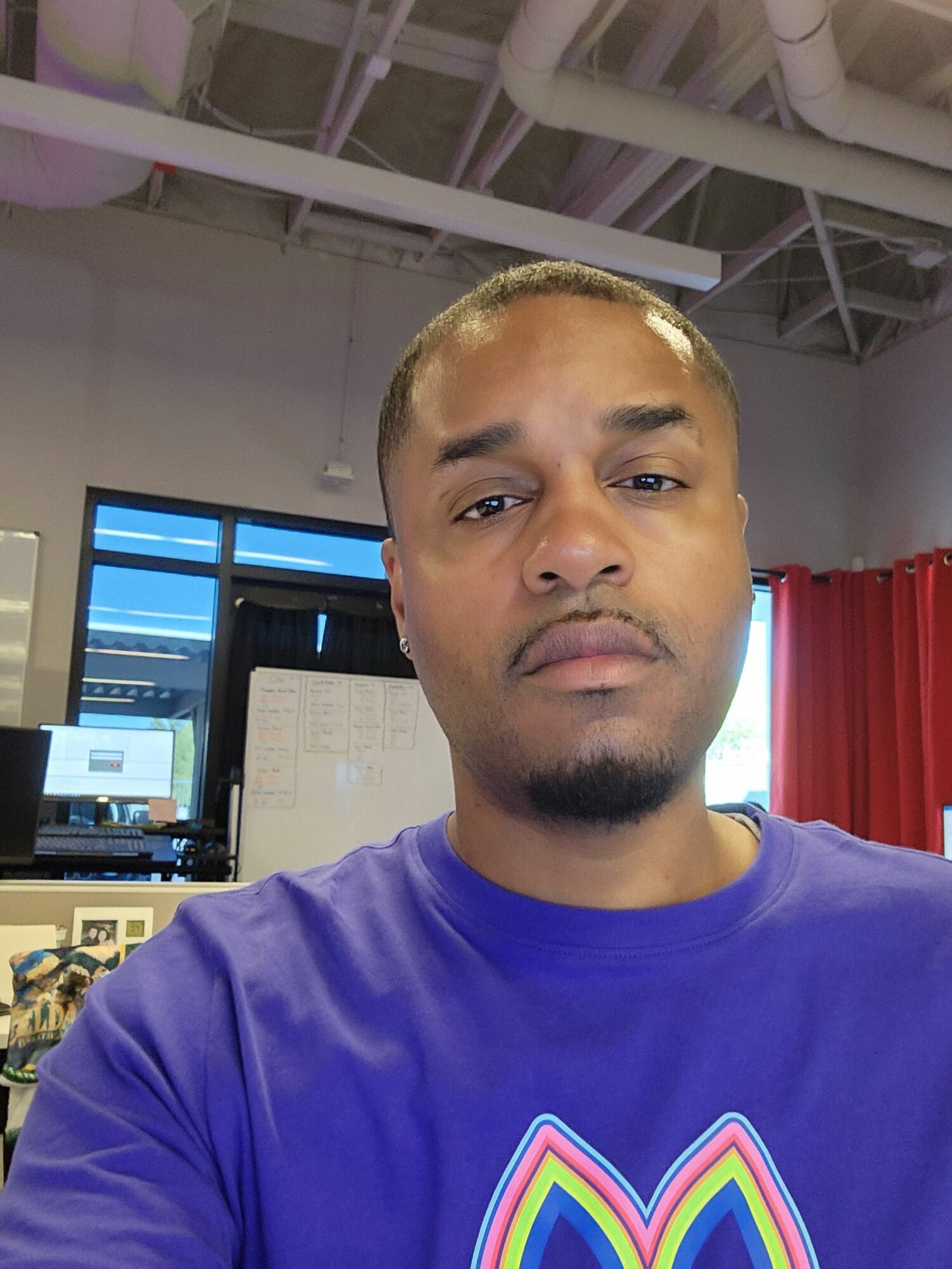 Young man with short hair and a goatee, wearing a purple shirt, smiling indoors with computer screens in background.