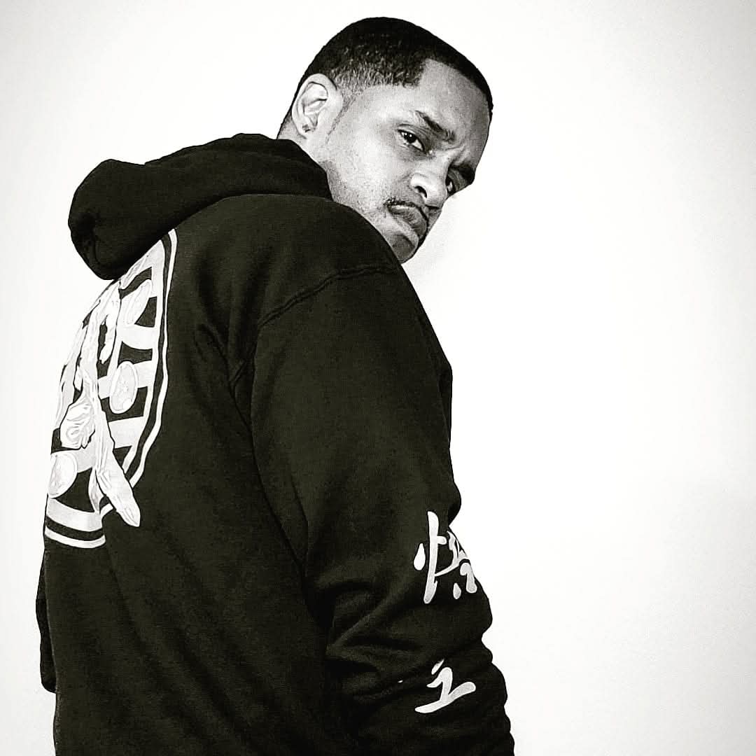 Person with short hair wearing a black hoodie, looking over shoulder, plain background.