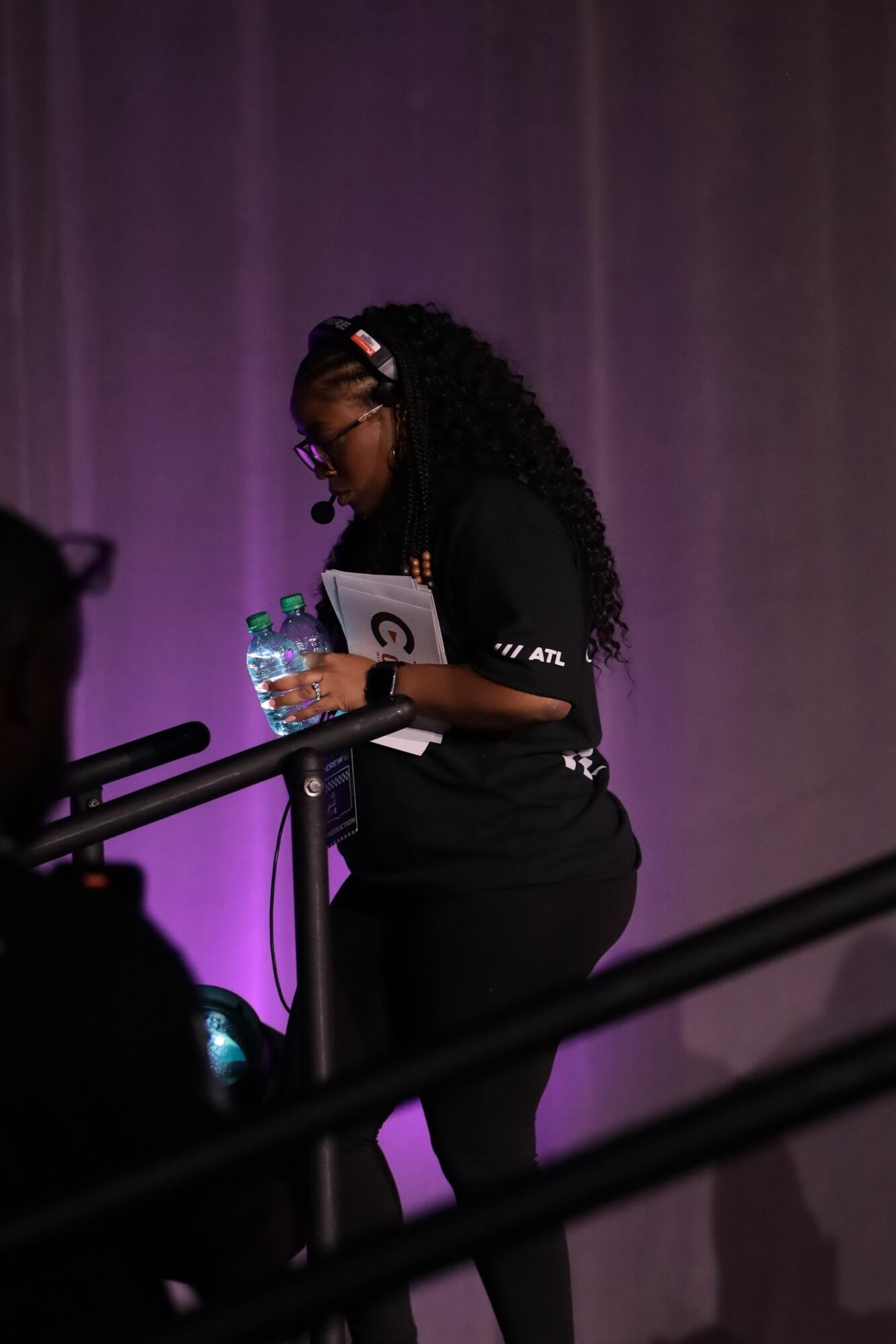 Person with braided hair holding a water bottle, sitting in front of a purple curtain, wearing dark clothing.