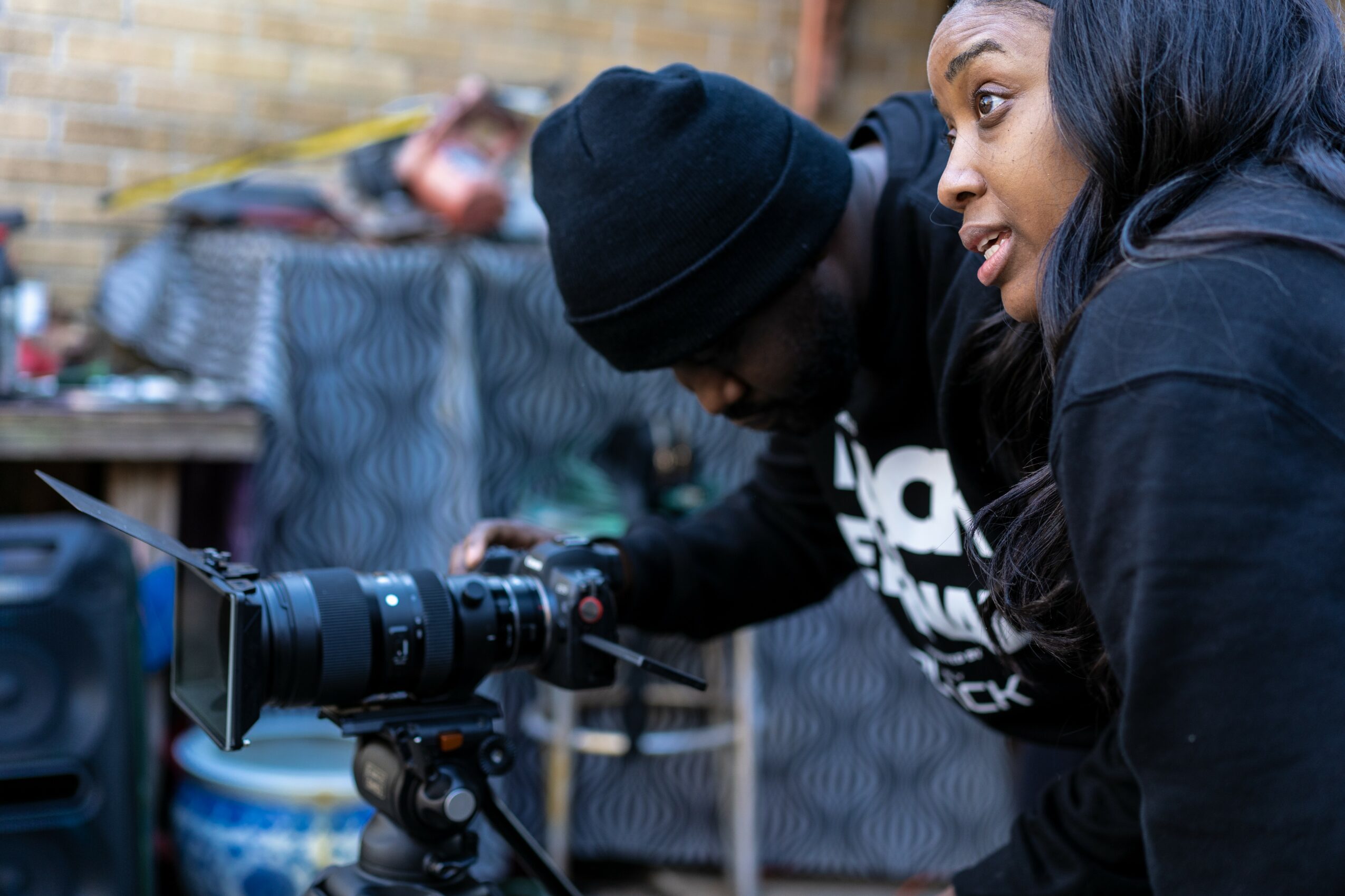Two people look at a camera mounted on a tripod outdoors, with a blurred background of a person and objects.
