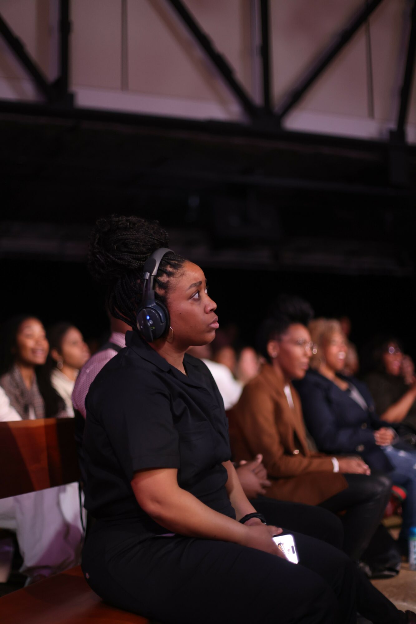 Woman with headphones leaning forward, surrounded by seated people in a dark indoor setting.