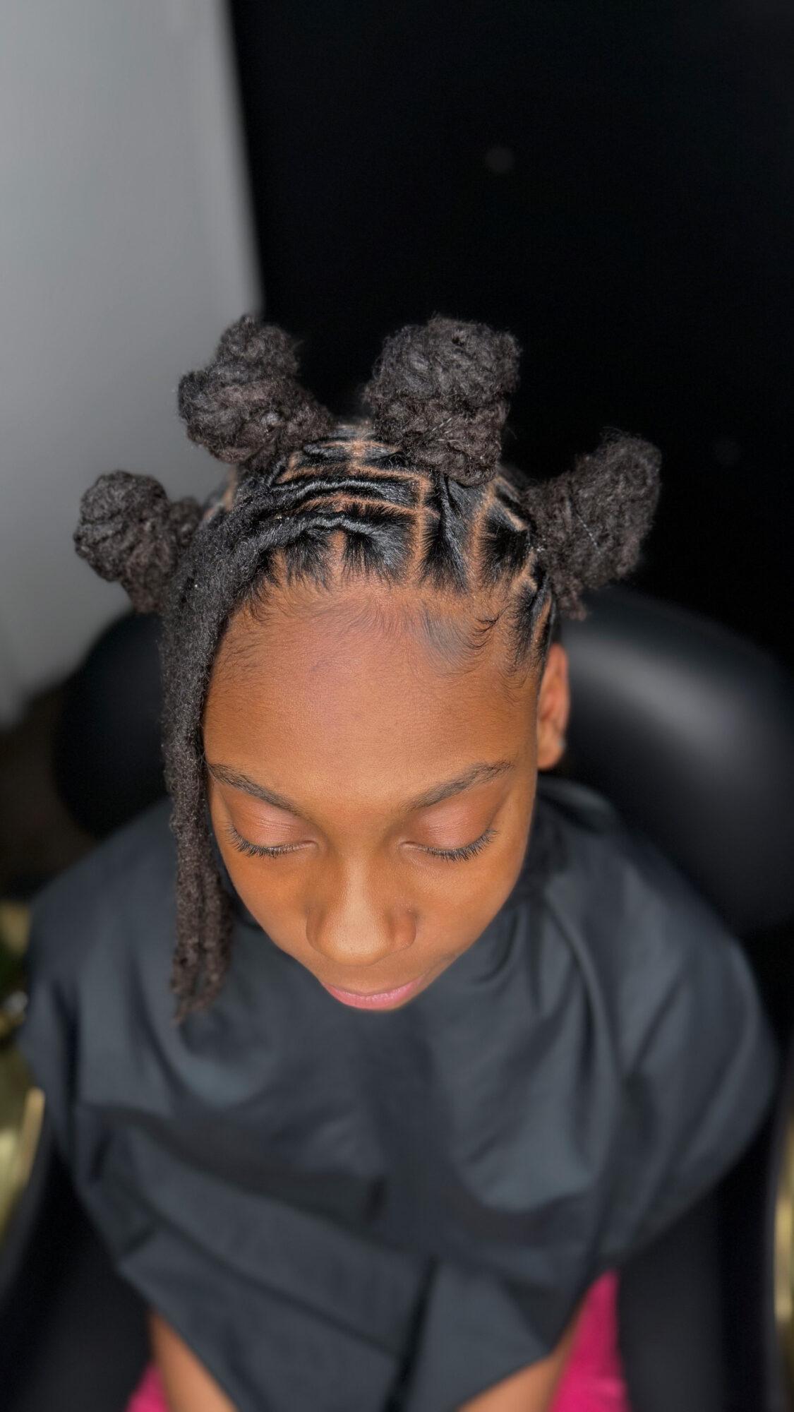 Person with styled hair in bantu knots, looking downward, wearing a black cape, indoors.