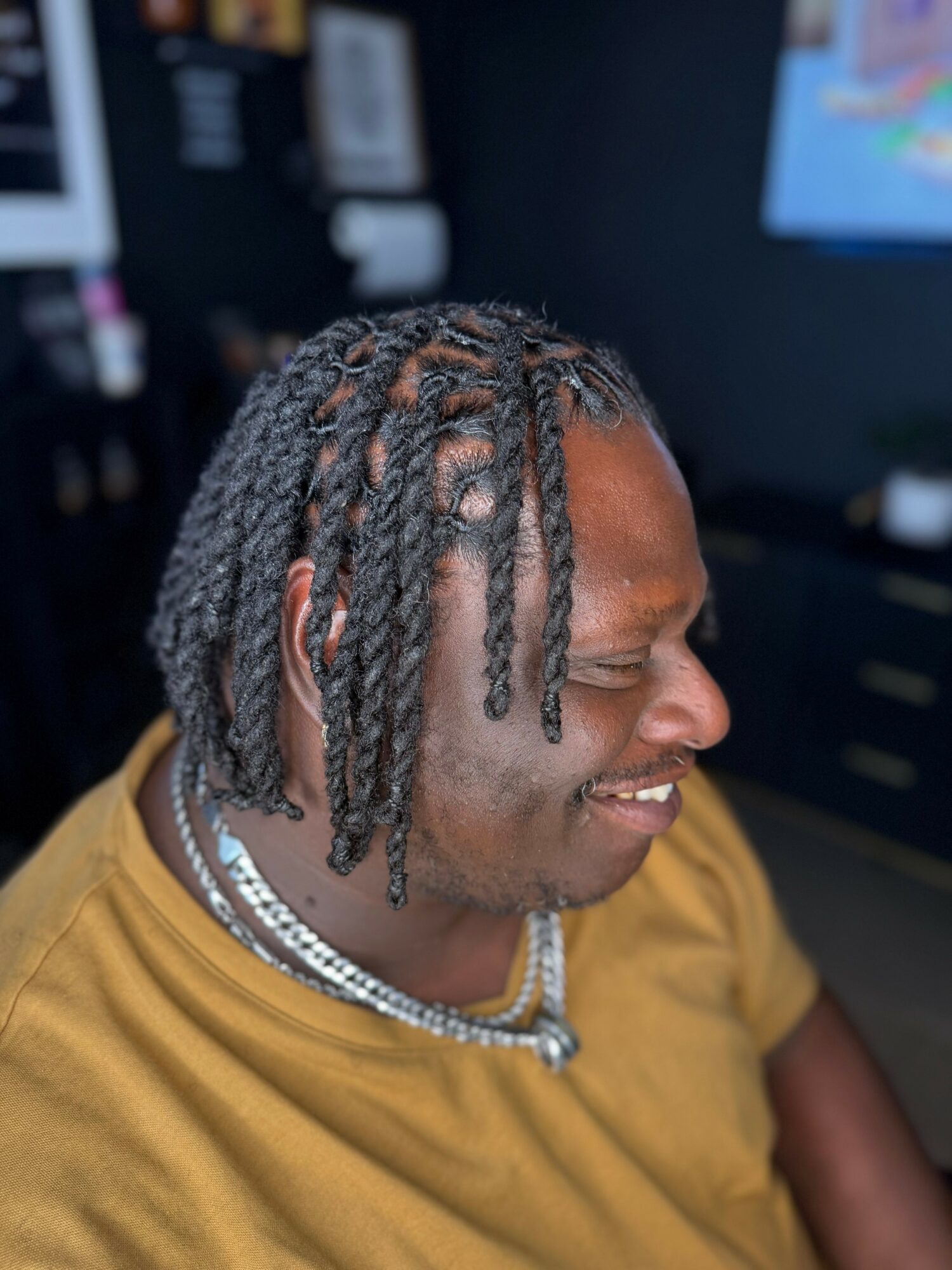 Man with styled dreadlocks wearing a mustard shirt and layered necklaces, smiling with eyes closed in indoor setting.