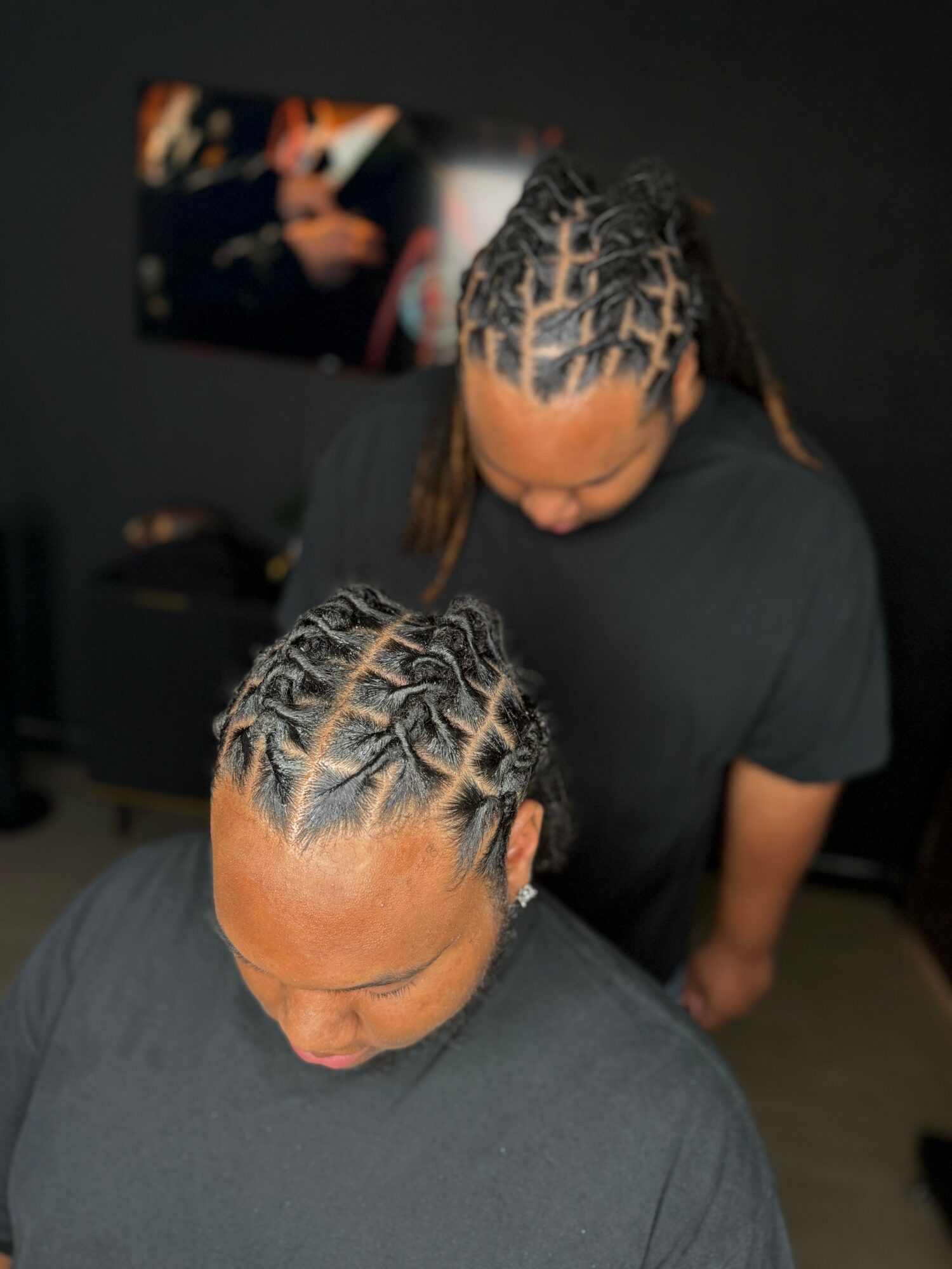 Two people with braided hairstyles, one in the foreground and one in the background, in a dark room.