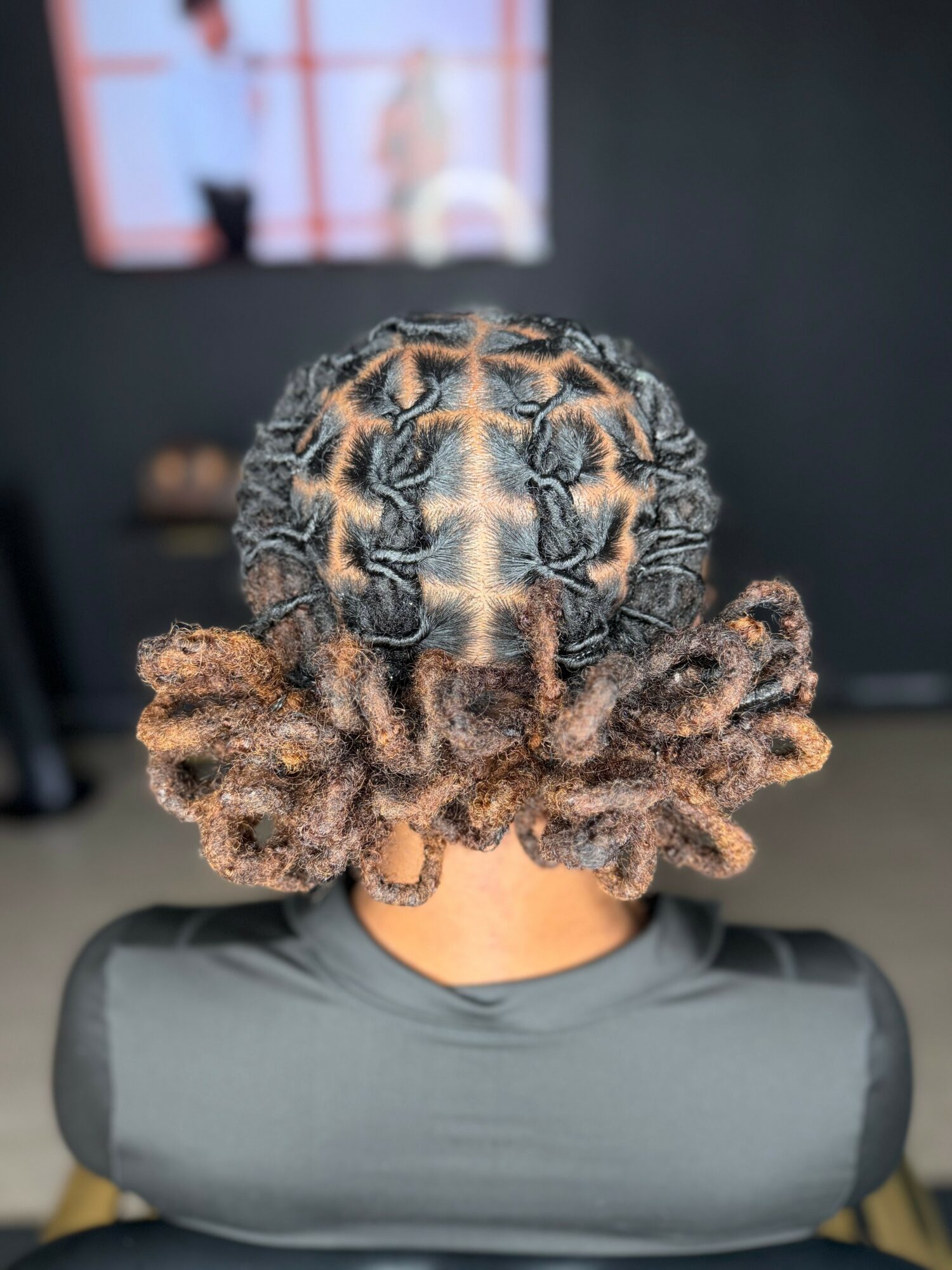 Back view of a person with intricately styled dreadlocks, sitting in a room with a dark wall and a TV screen.