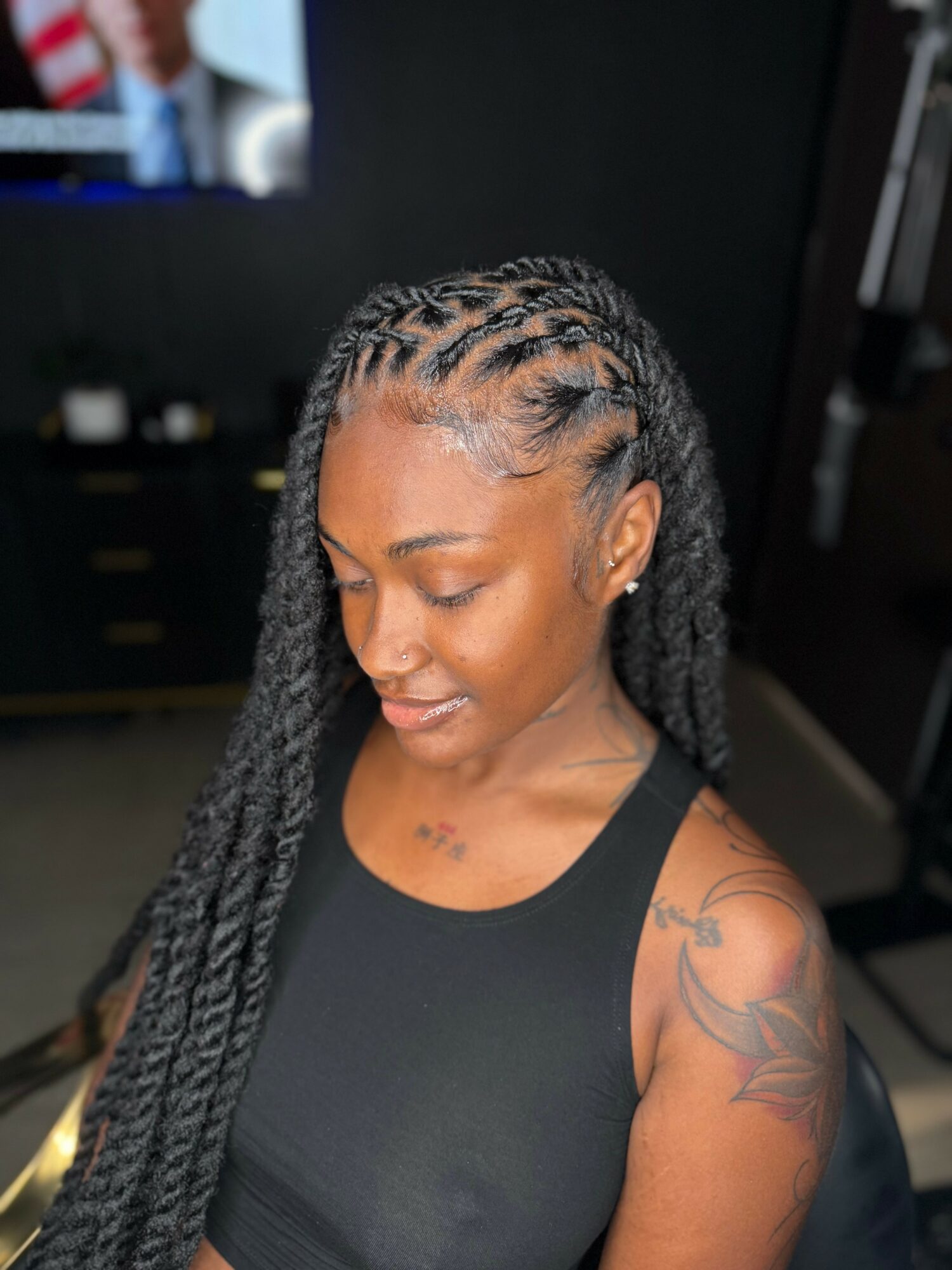 Young woman with braided hairstyle looking down, wearing a black sleeveless top, tattoo on her shoulder, in a room.