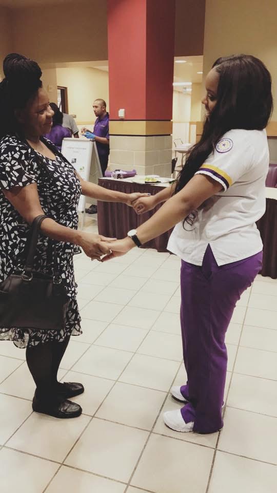Two women holding hands and facing each other in a shopping mall, with people and a red pillar in the background.