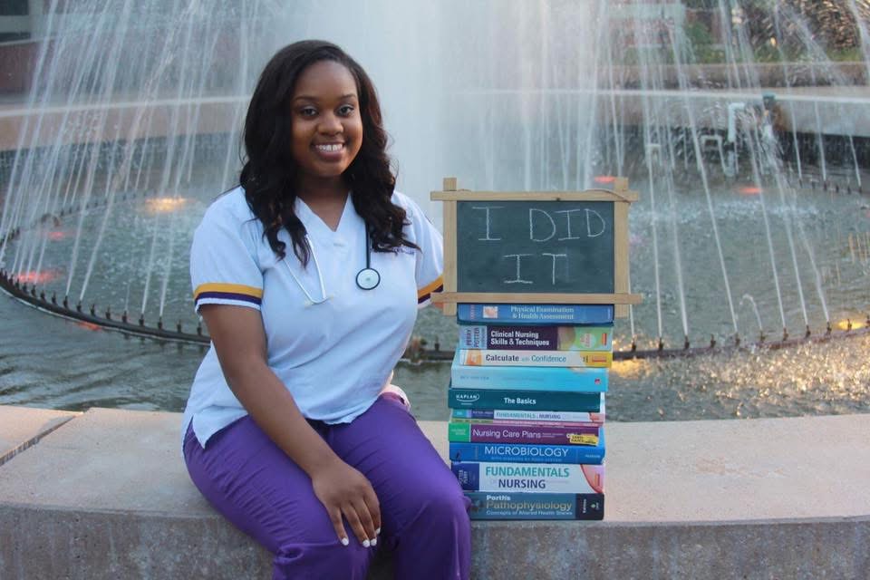 Young woman sitting by a fountain with a stack of books and a chalkboard sign that says 'I DID IT'.