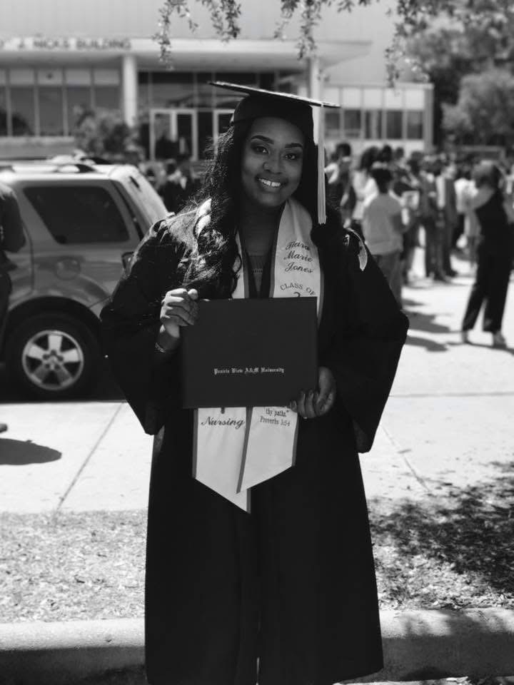 Graduate in cap and gown holding diploma outdoors with people and cars in background.