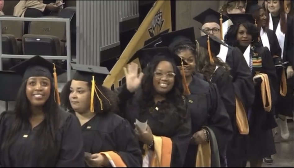 Group of graduates in caps and gowns walking in a line, smiling and waving, at a graduation ceremony.