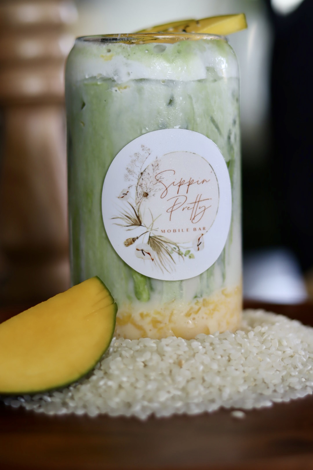 Tall glass with green beverage, lemon slice on top, label with floral design, placed on a textured surface with mango and rice nearby.