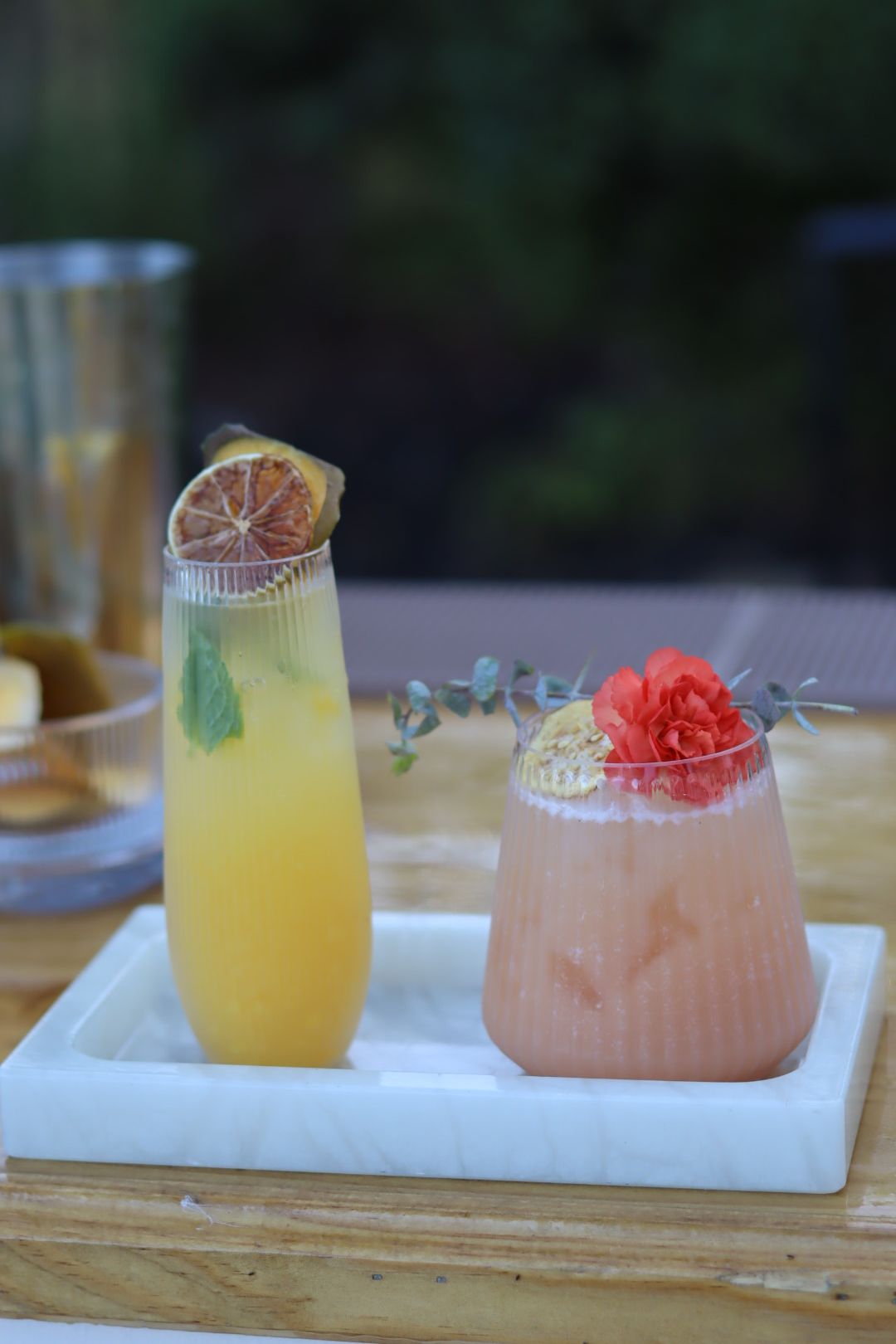 Two colorful drinks on a white tray, one yellow with a lime slice, the other pink with a red flower garnish, outdoors.