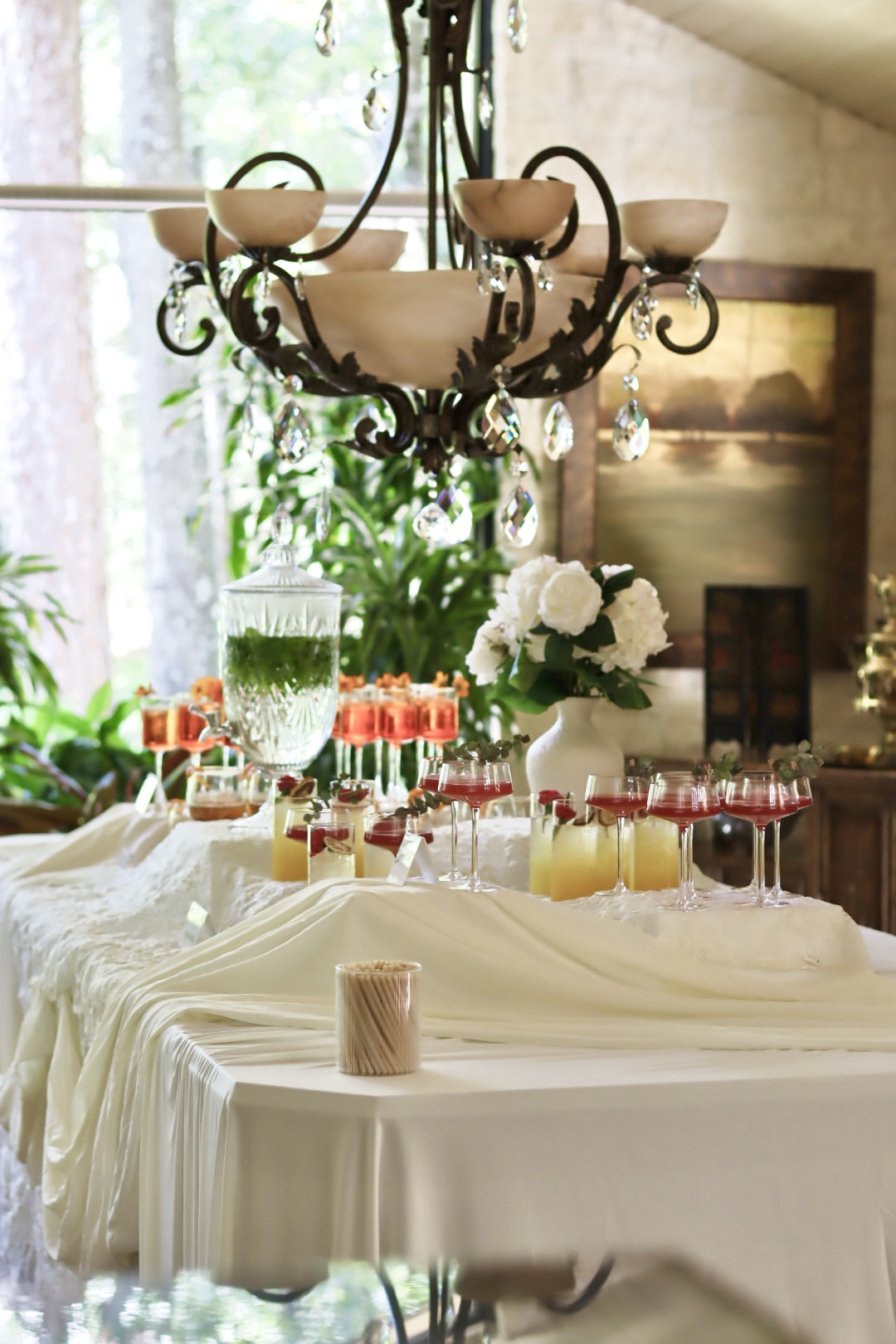 Elegant dining table with white tablecloth, floral centerpiece, and chandelier overhead in a bright room.