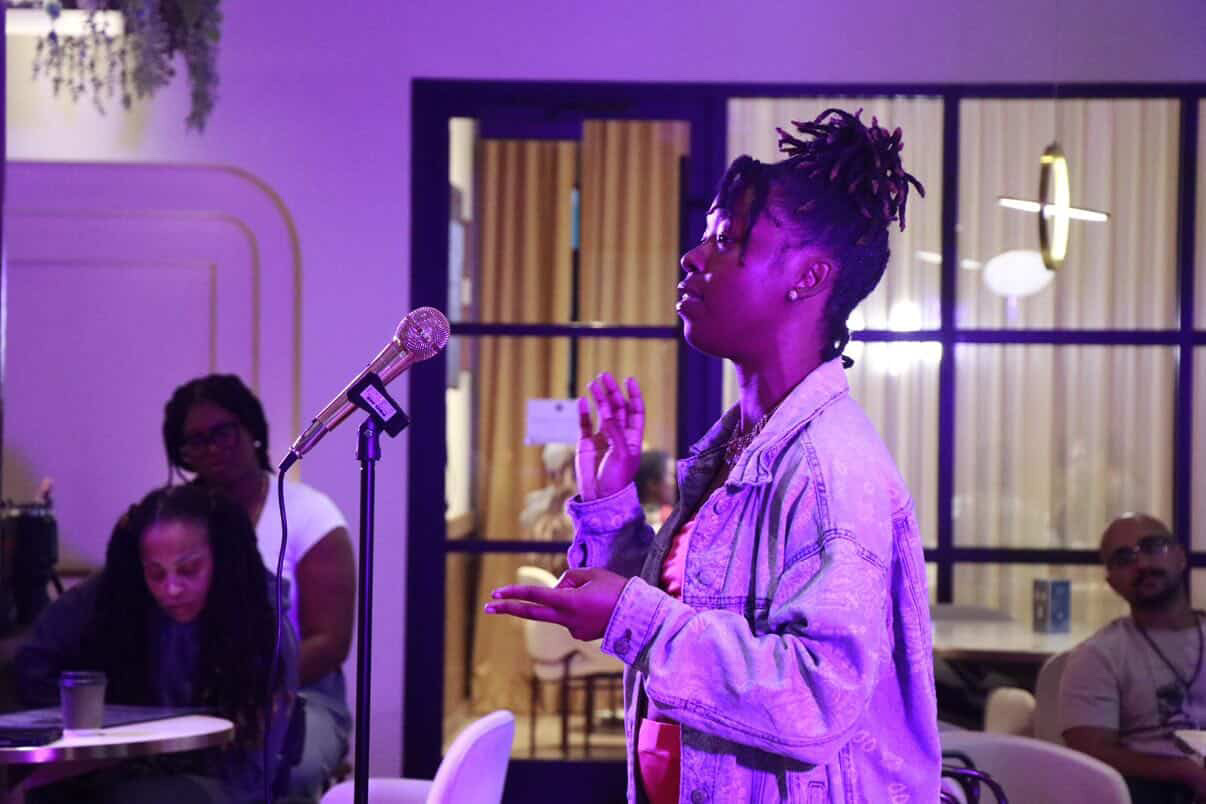 Person with dreadlocks speaking into a microphone, gesturing with hands, in a room with seated audience members, purple lighting.
