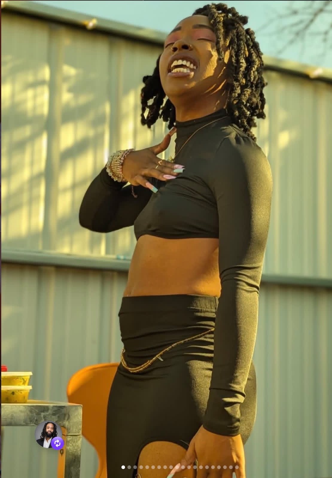 Young woman with dreadlocks smiling, wearing a black crop top and skirt, standing outdoors near a fence.