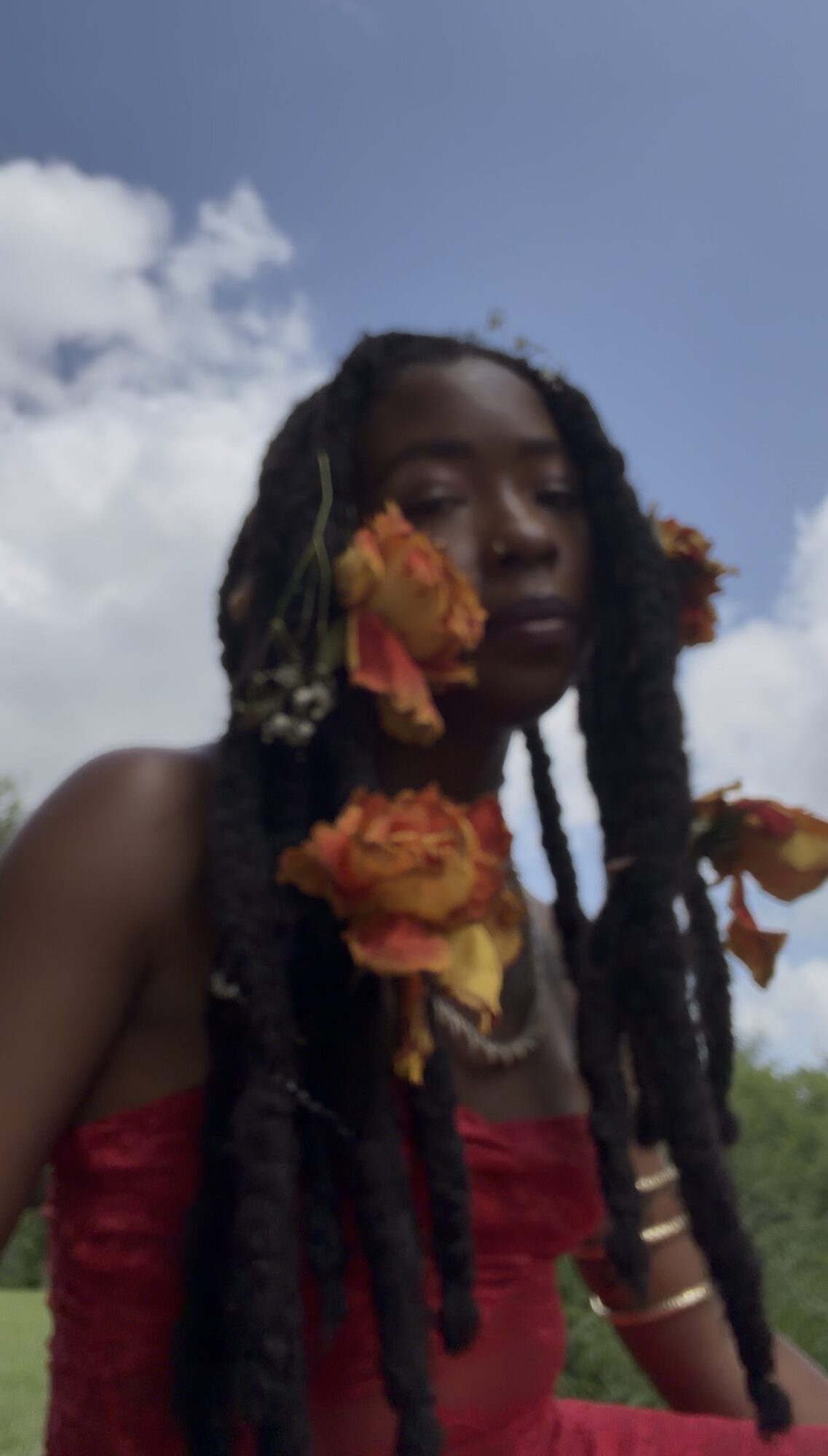Person with long braided hair outdoors, flowers in hair, wearing a red top, sky and clouds in background.