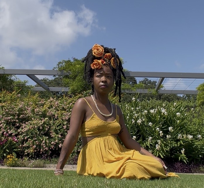 Woman sitting on grass in yellow dress with floral headpiece, garden background, blue sky, clouds.