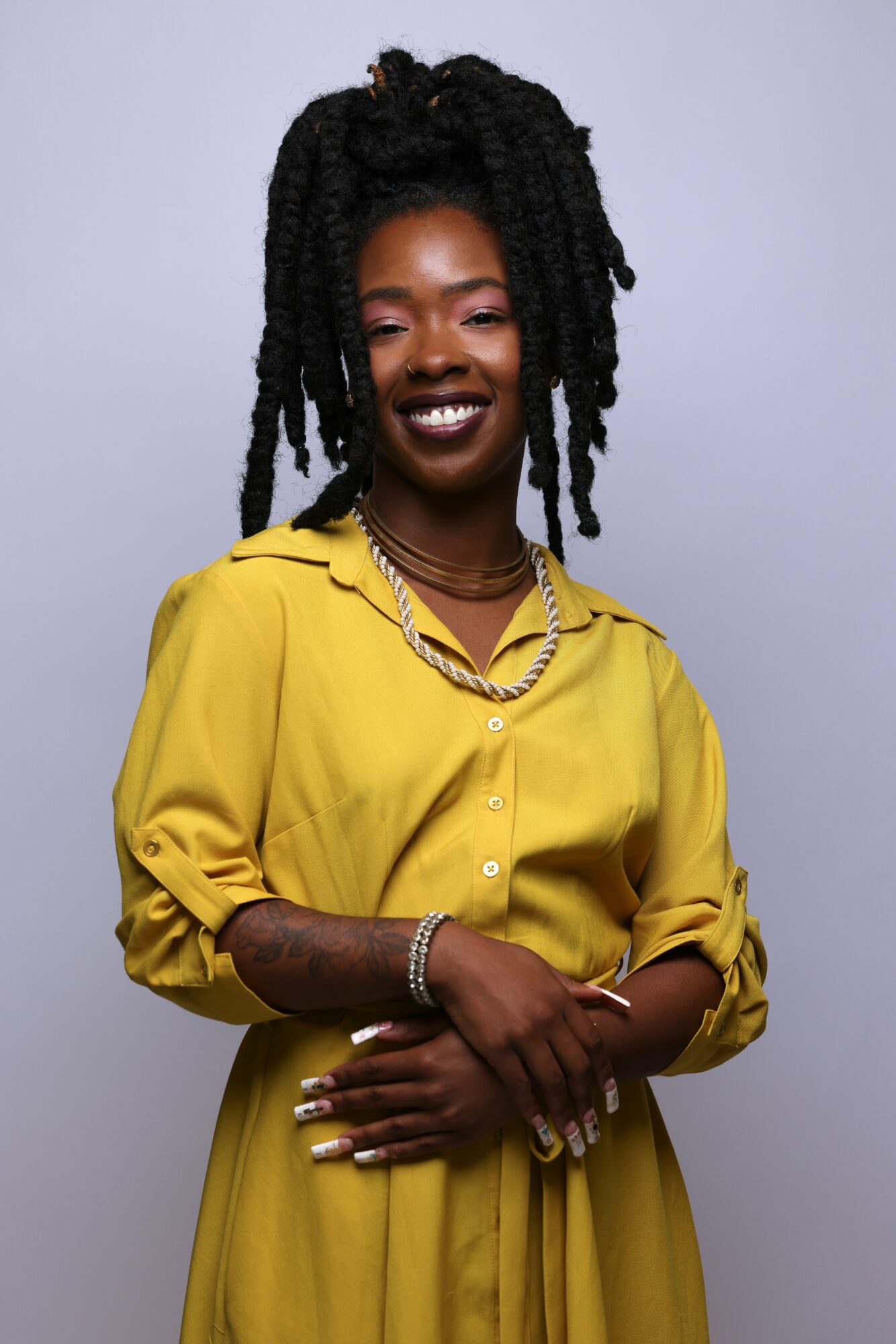 Smiling woman with long twisted hair wearing a yellow shirt and jewelry, standing against a plain background.