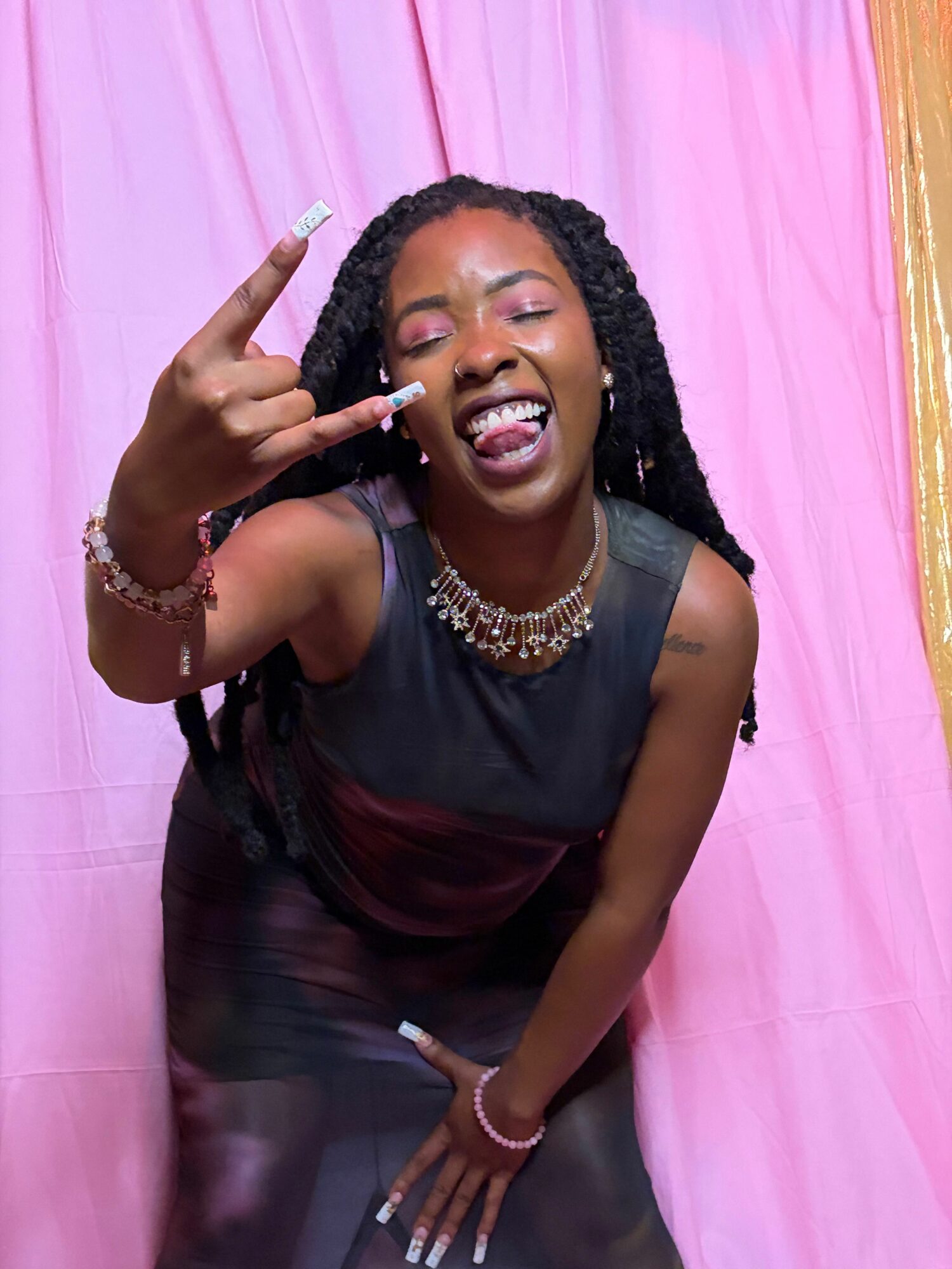 Woman lying on pink surface, smiling, making a hand gesture, wearing jewelry and dark clothing.