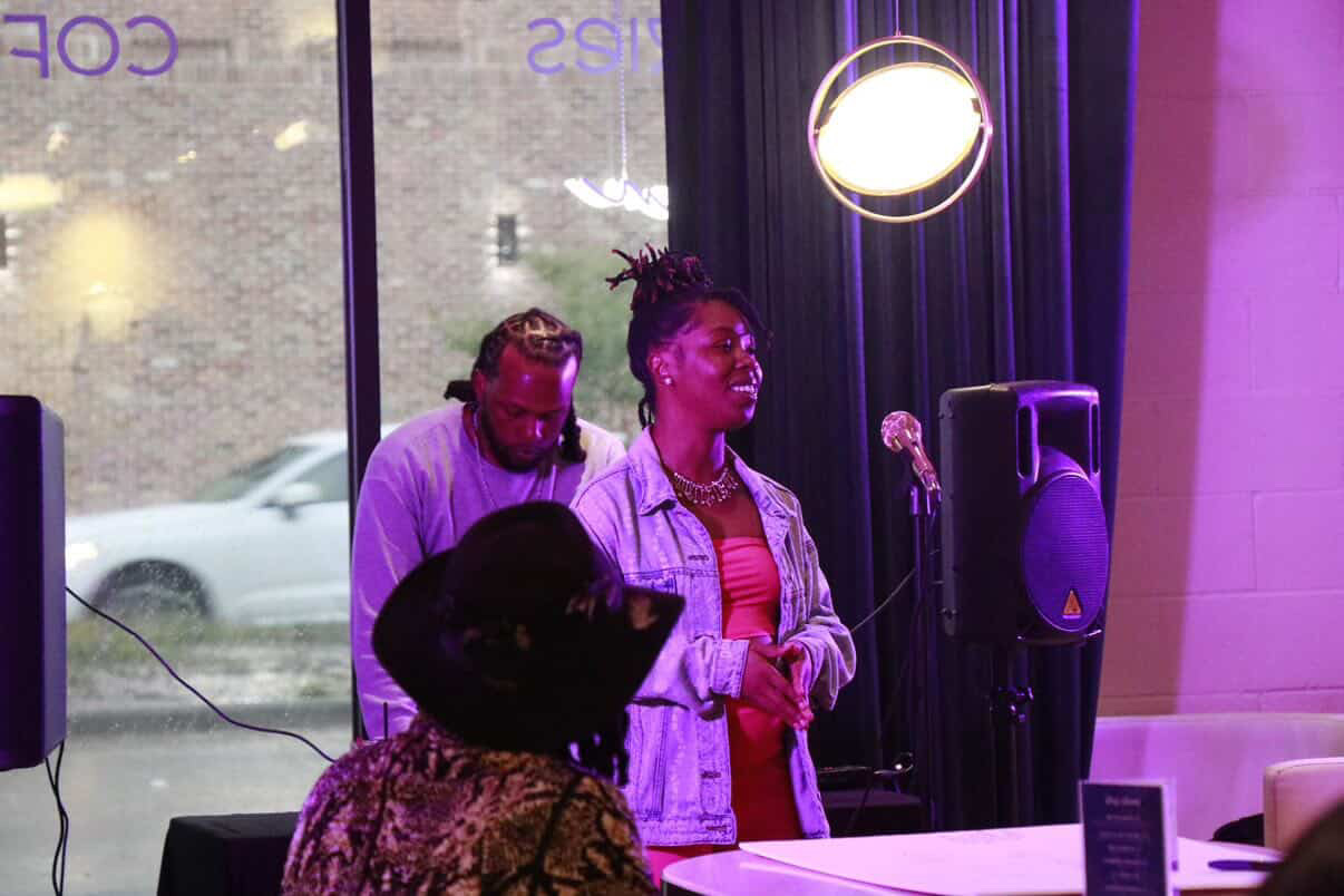 Two performers on stage, one singing with eyes closed, the other playing an instrument, with a microphone and speakers nearby.