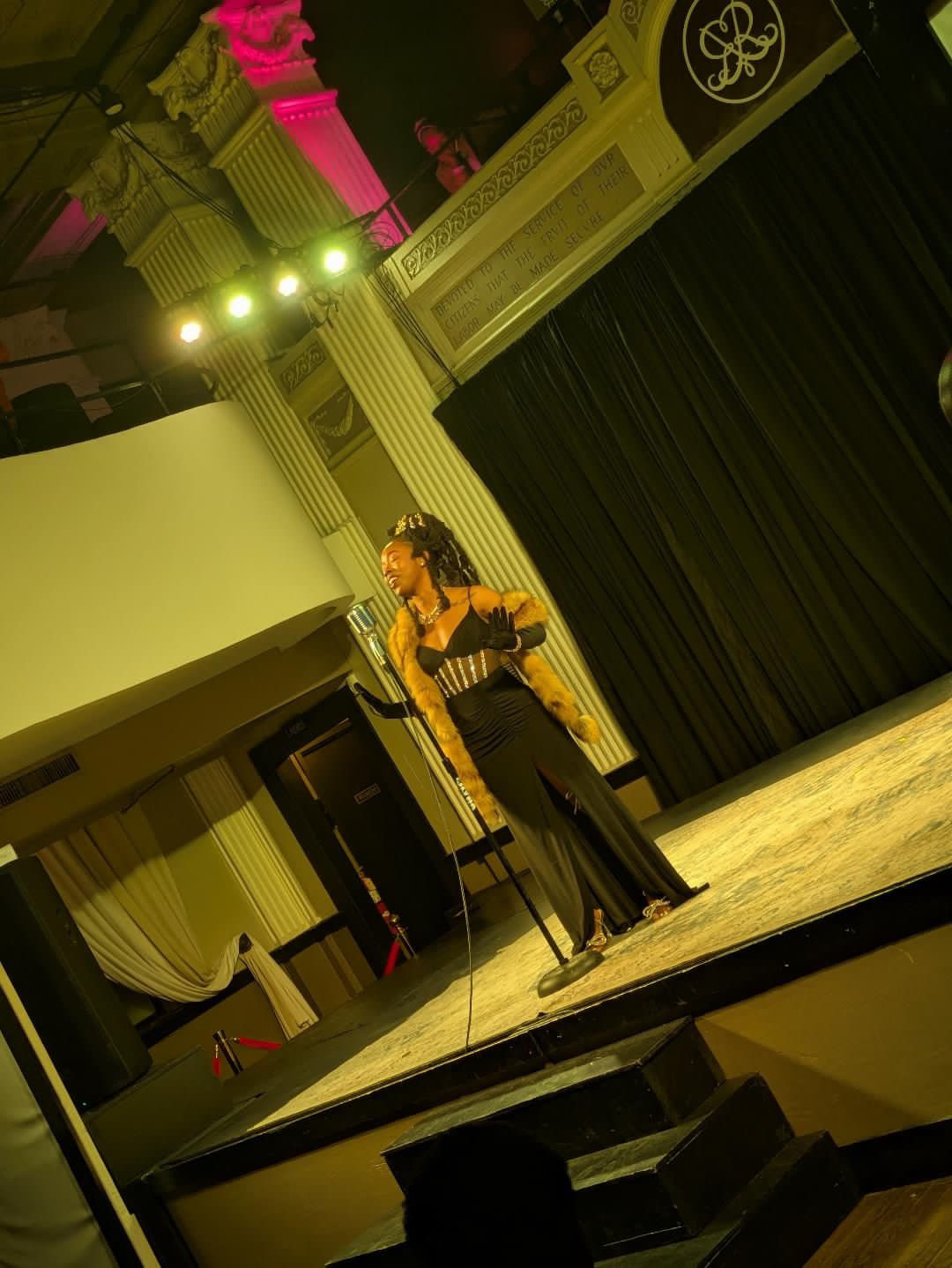 Performer on stage with a microphone, black dress, in a decorated theater with columns and curtains.