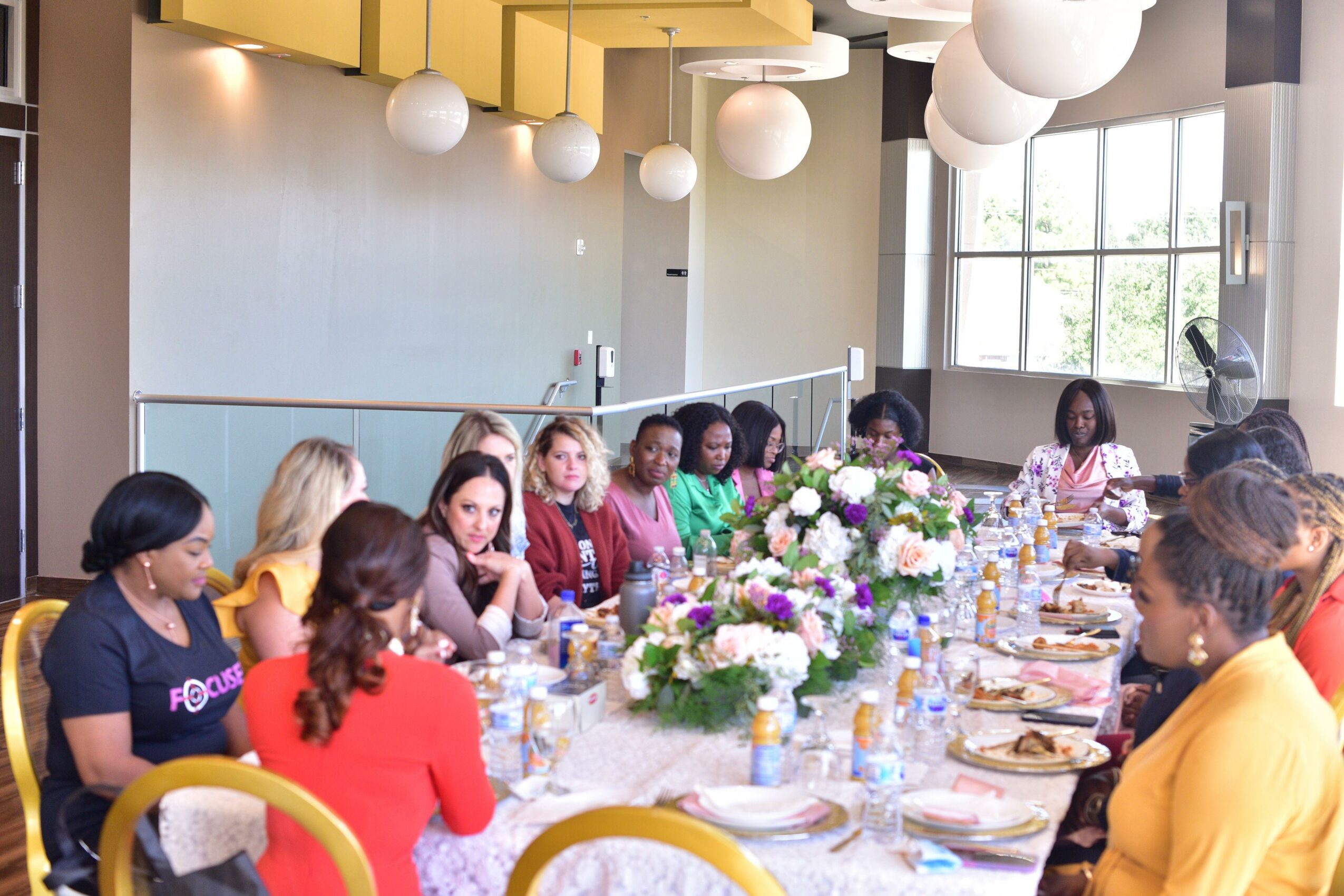 Group of women sitting around a long dining table with floral centerpieces, in a bright room with large windows and hanging lights.