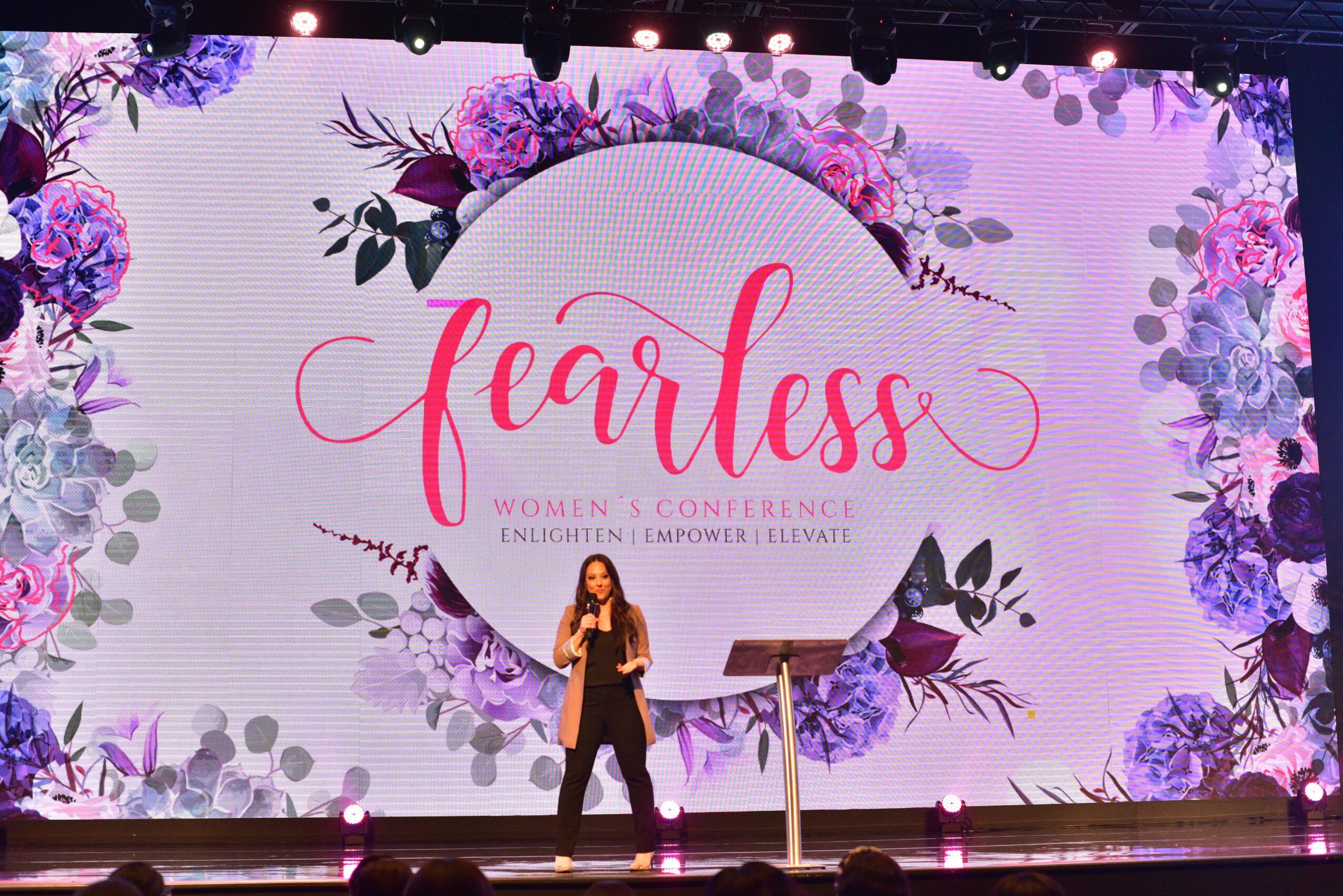 Woman speaking on stage at women’s conference with floral background and large 'fearless' text.
