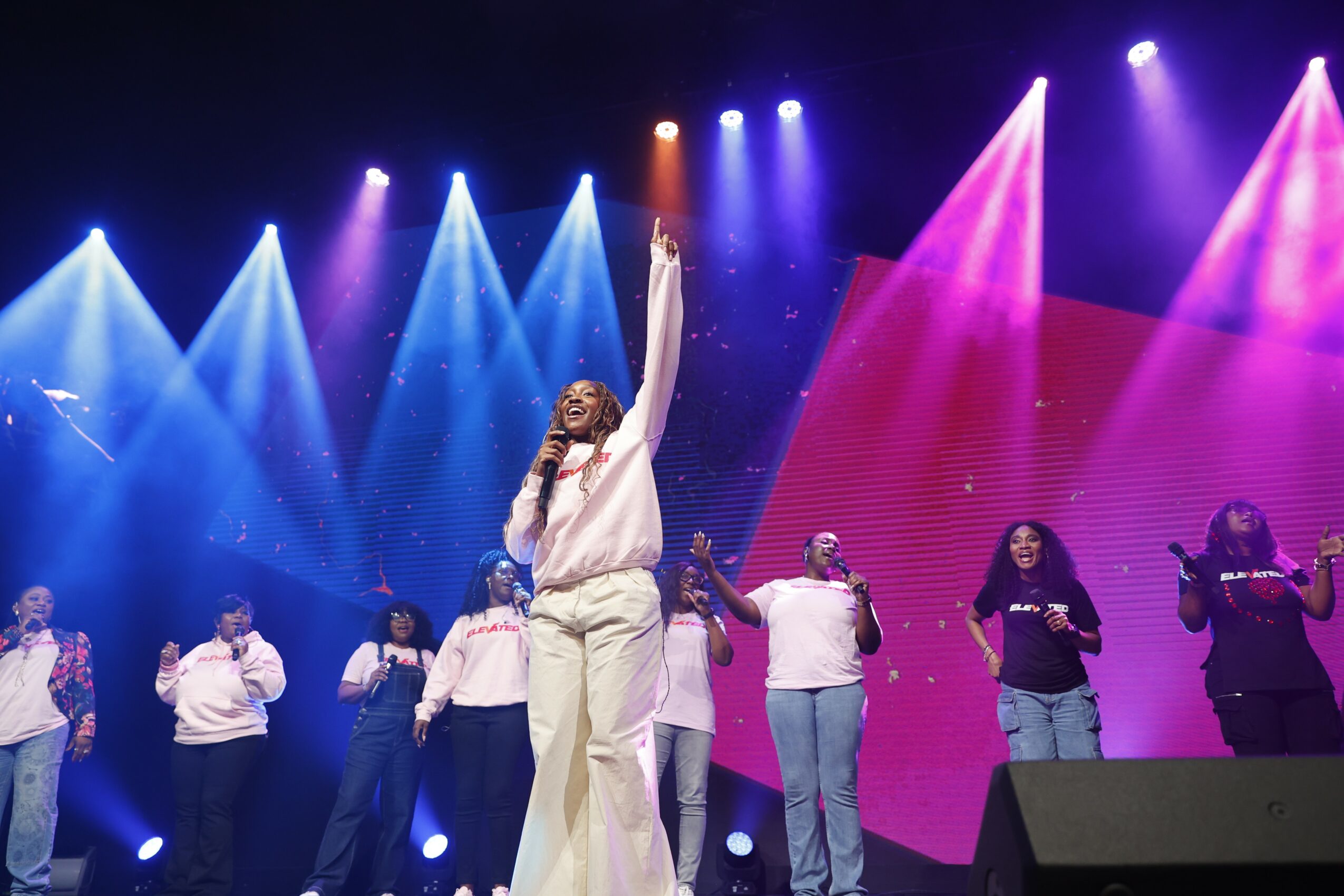 Group of people on stage with colorful lights, one person raising an arm, others standing and singing.