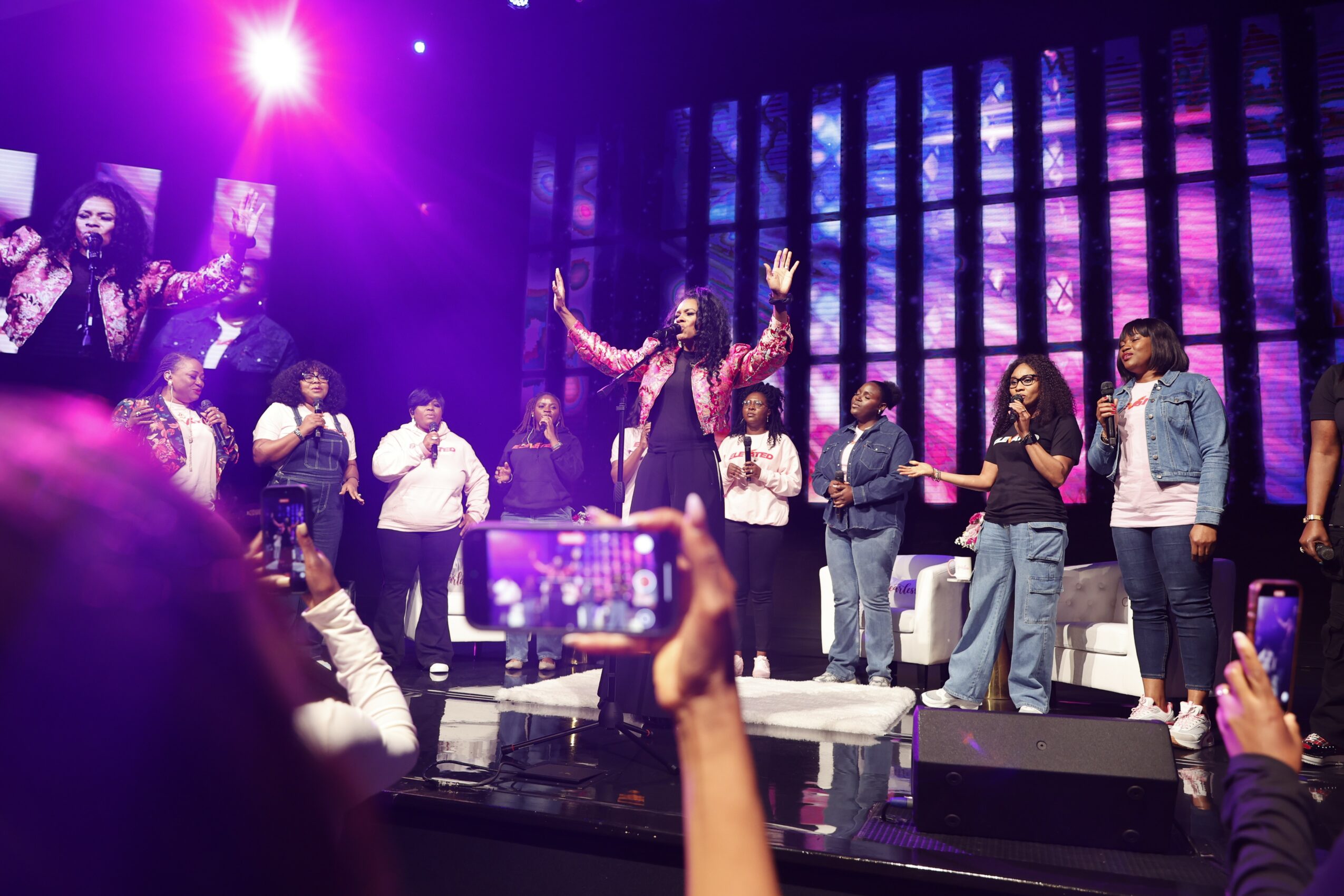 Group of people on stage with a performer in the center, colorful lighting, and audience members taking photos.
