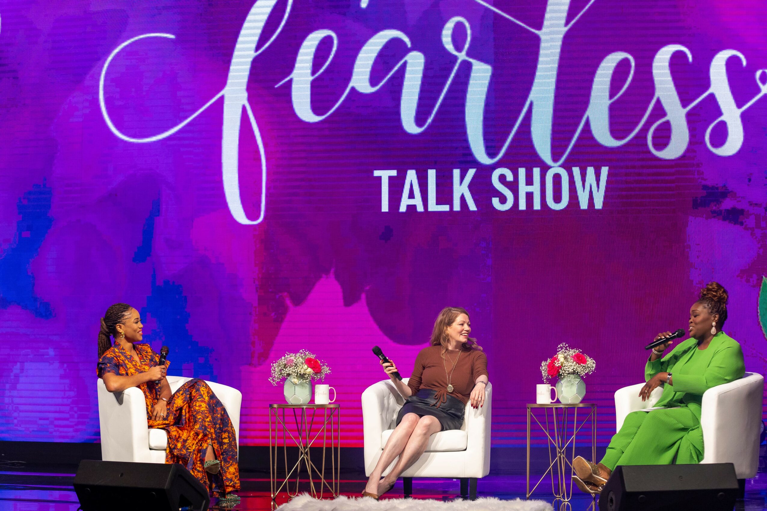 Three women sit on stage in white chairs with small tables and flowers, with a purple background and large text reading 'fearless TALK SHOW'.