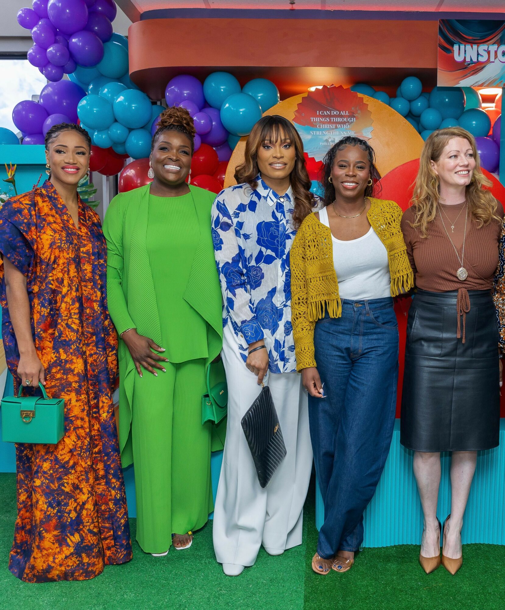 Five women standing together in front of colorful balloons and a backdrop, smiling and posing for the photo.