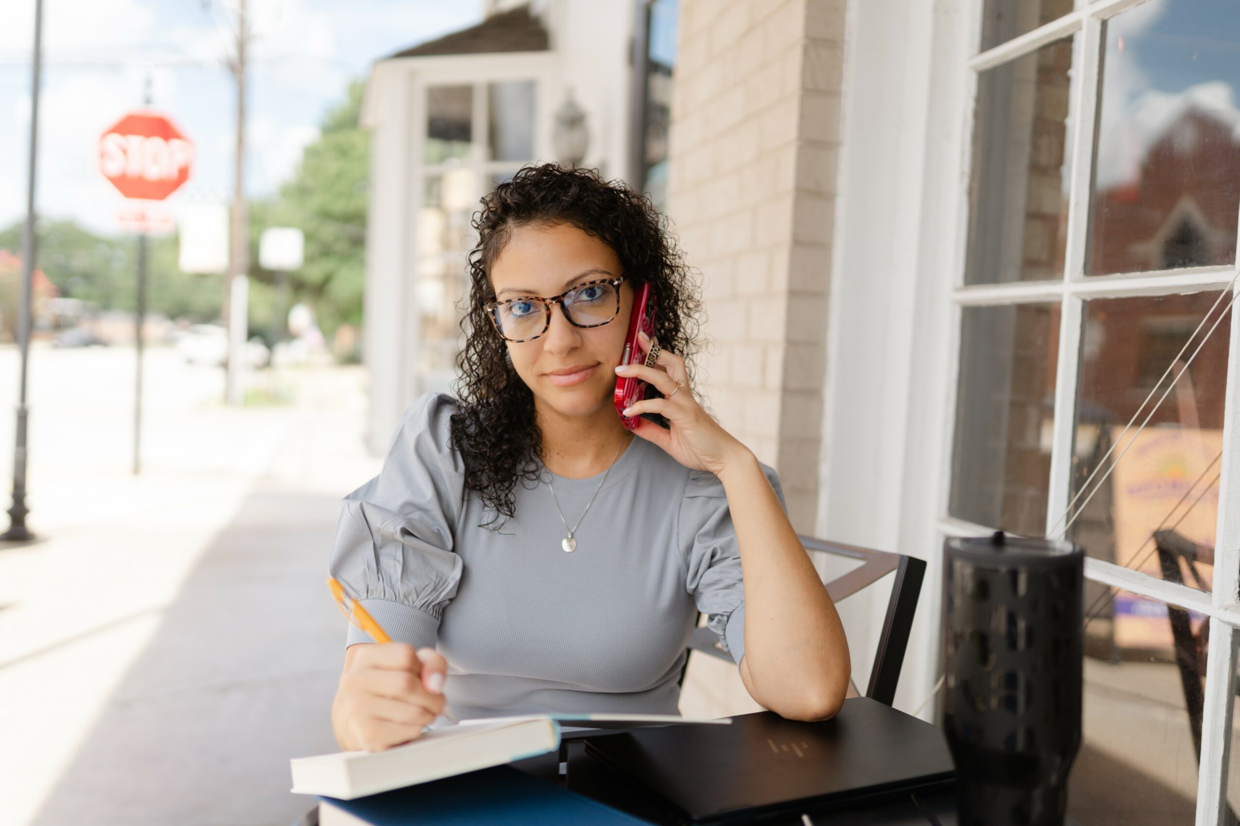 Woman with glasses sitting at a table outside, talking on phone, holding a pen, with a notebook and black container on table.