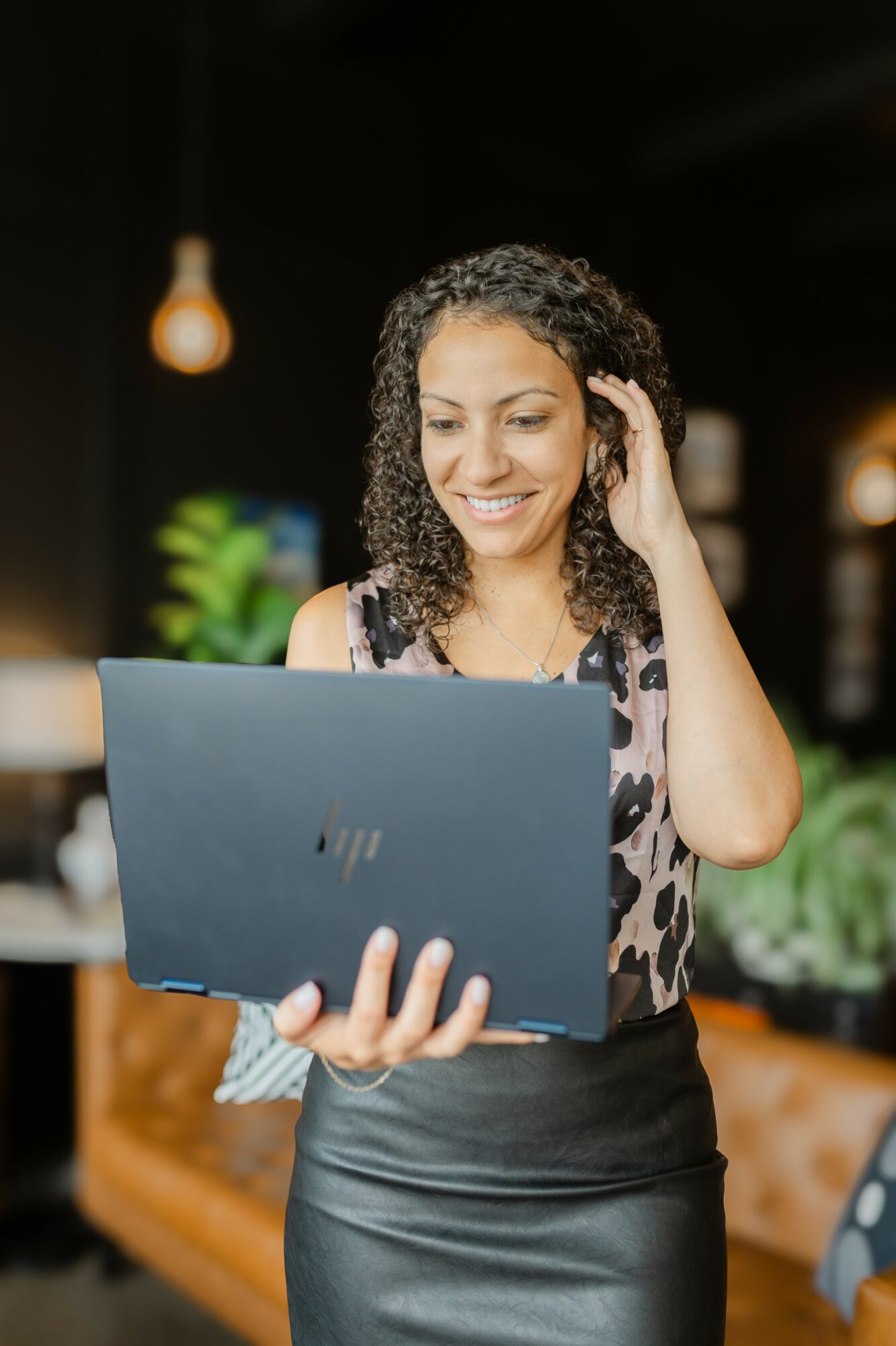 Woman with curly hair smiling while looking at a laptop in a cozy indoor setting.