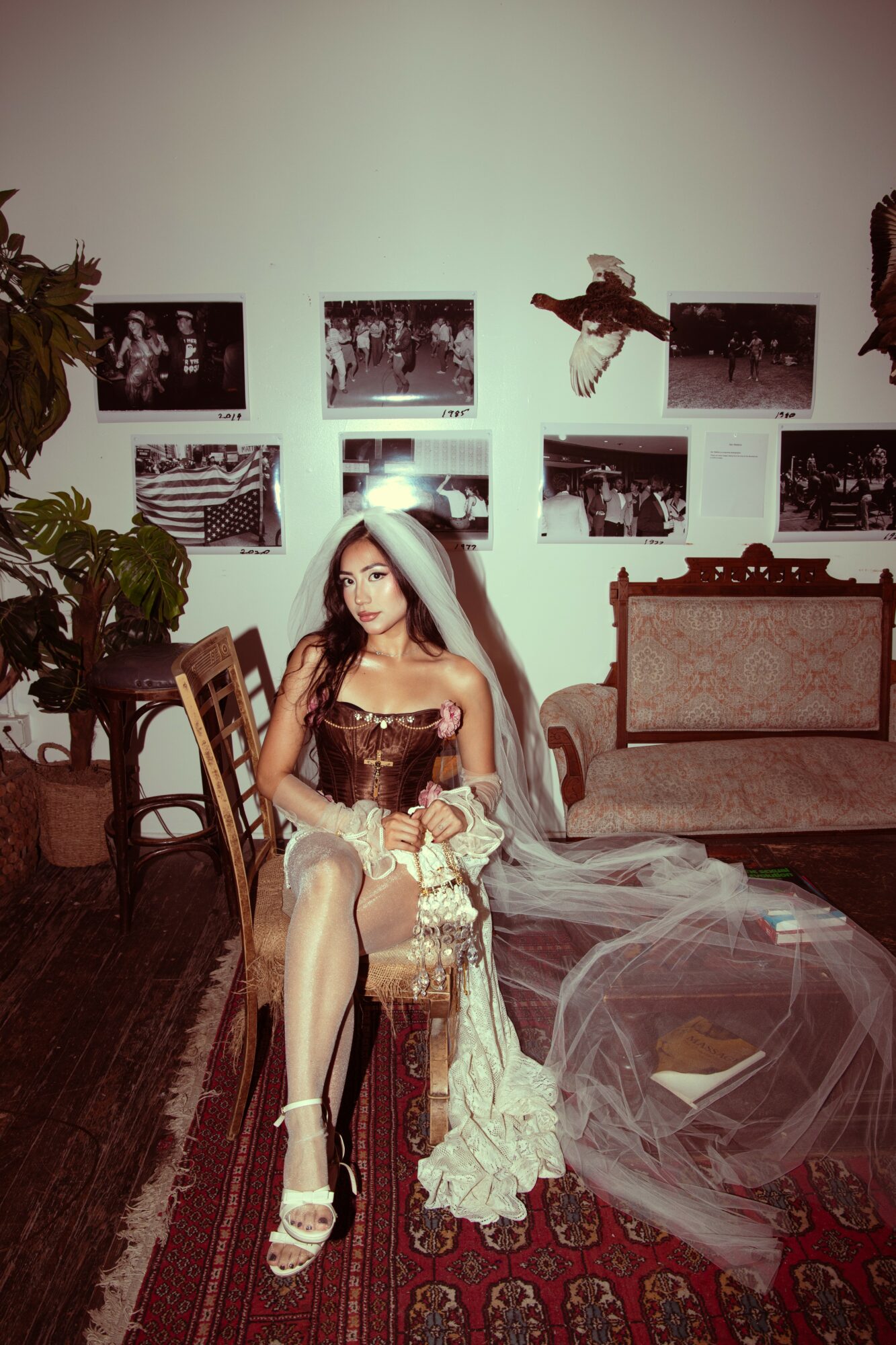 Young woman in vintage dress with veil sitting on a chair in a decorated room, with a sofa and framed pictures on the wall.