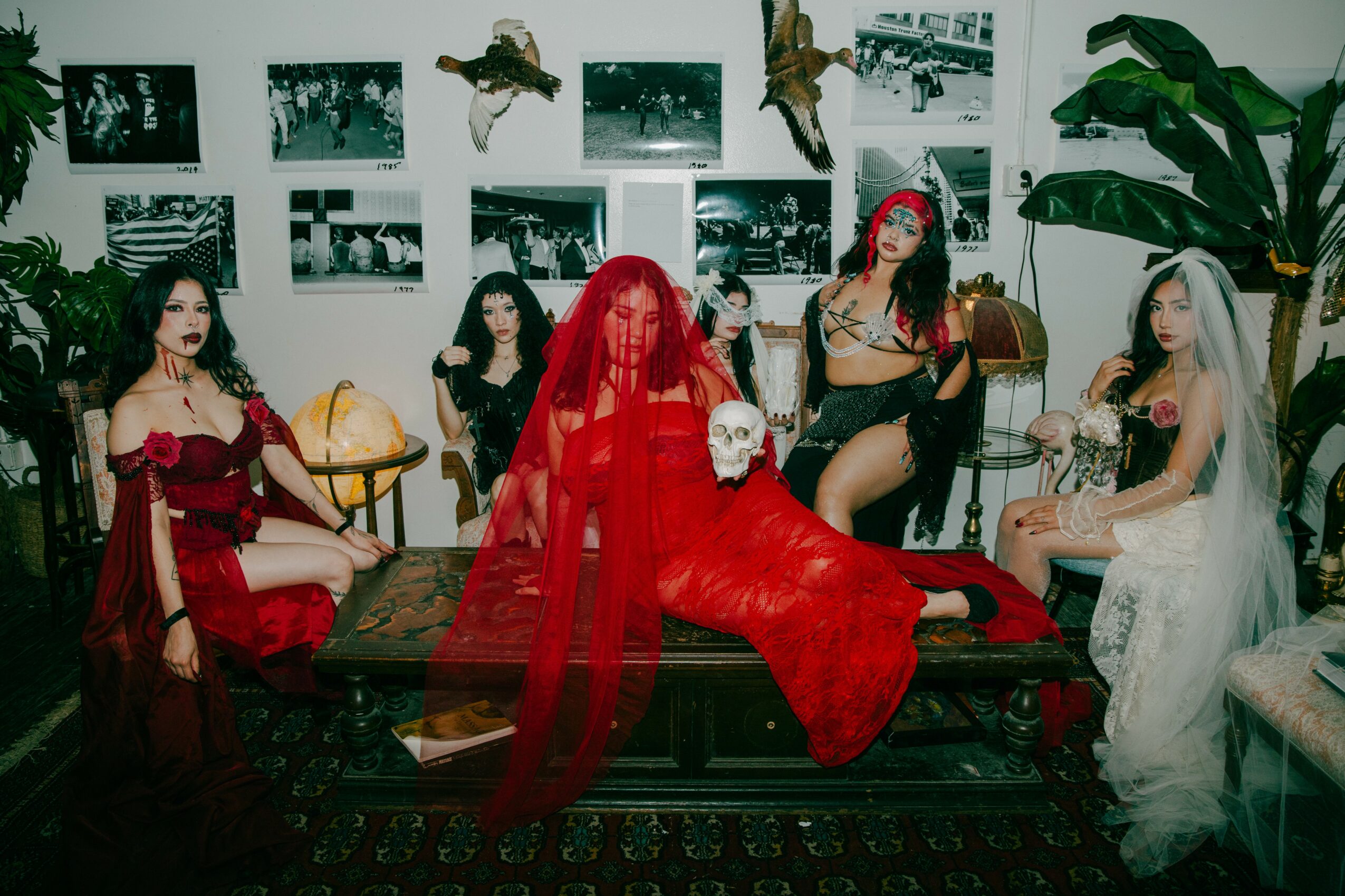 Group of women in costumes sitting and standing in a decorated room with photos and plants on the wall.