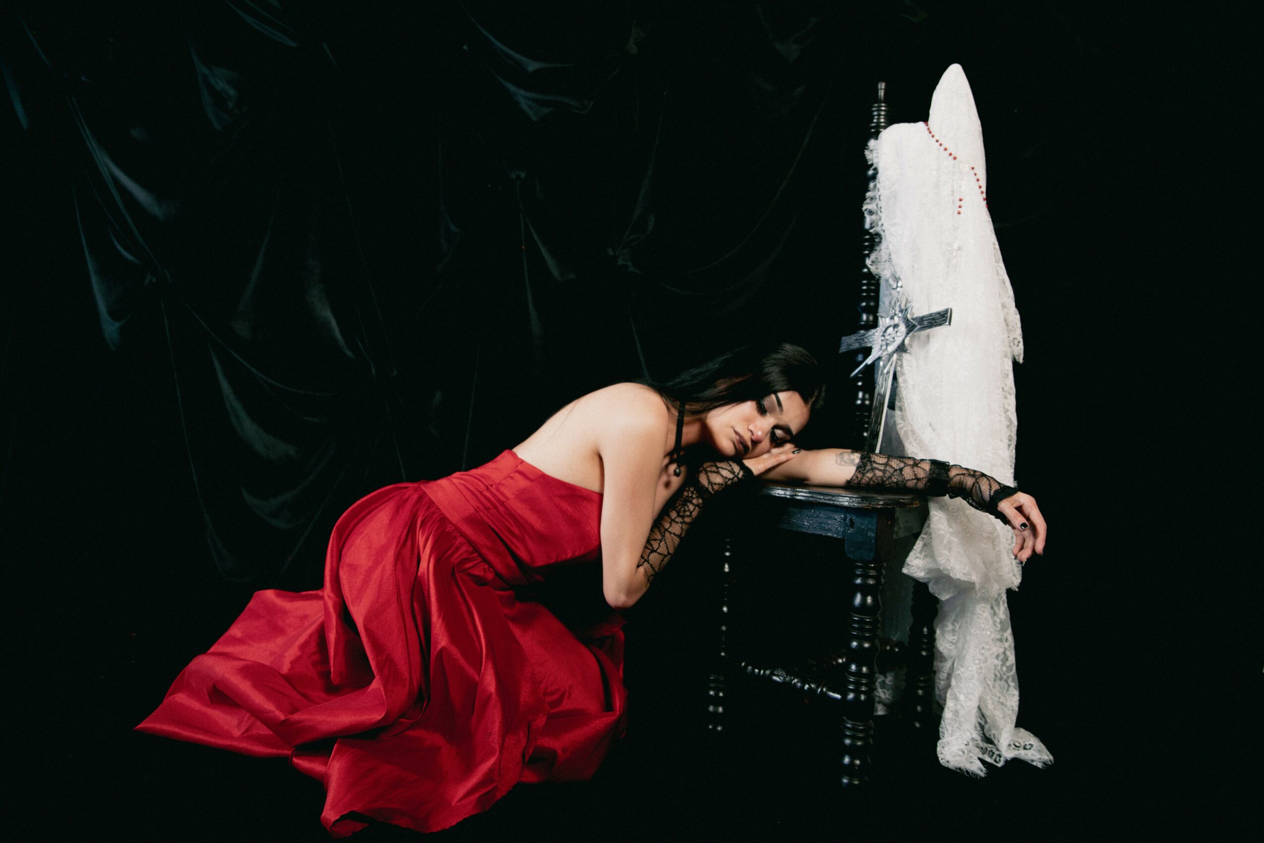 Woman in red dress leaning on a table with a white dress on a mannequin, against a black background.