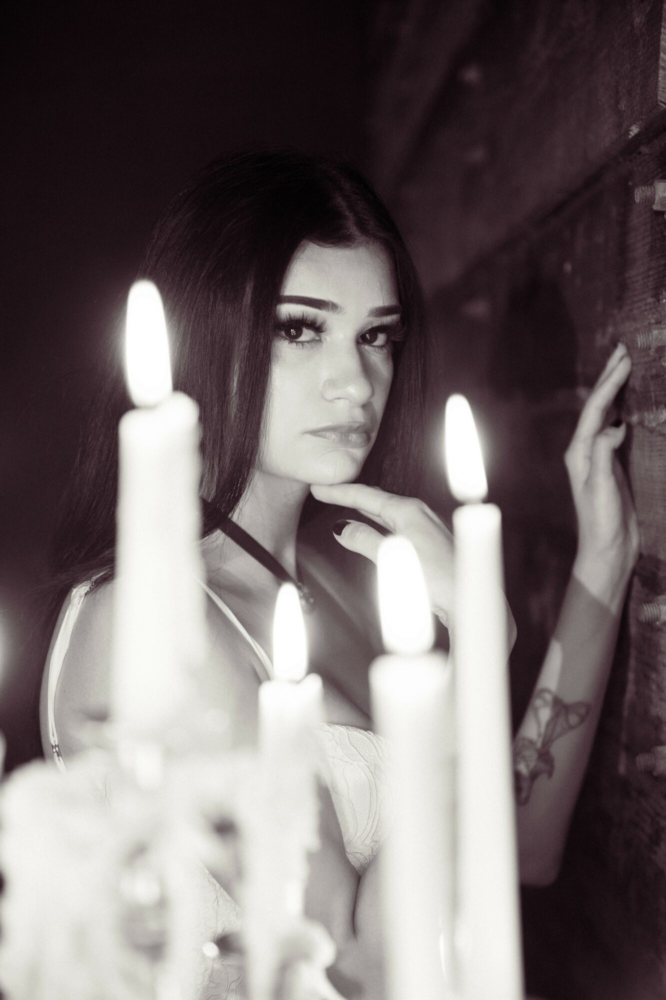 Young woman with dark hair and makeup surrounded by lit candles, looking at the camera, against a dark background.