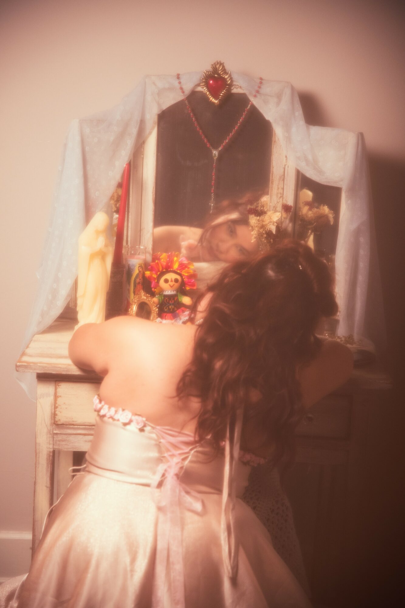 A woman in a white dress with dark hair faces a shrine with a framed picture, jewelry, and decorative items.