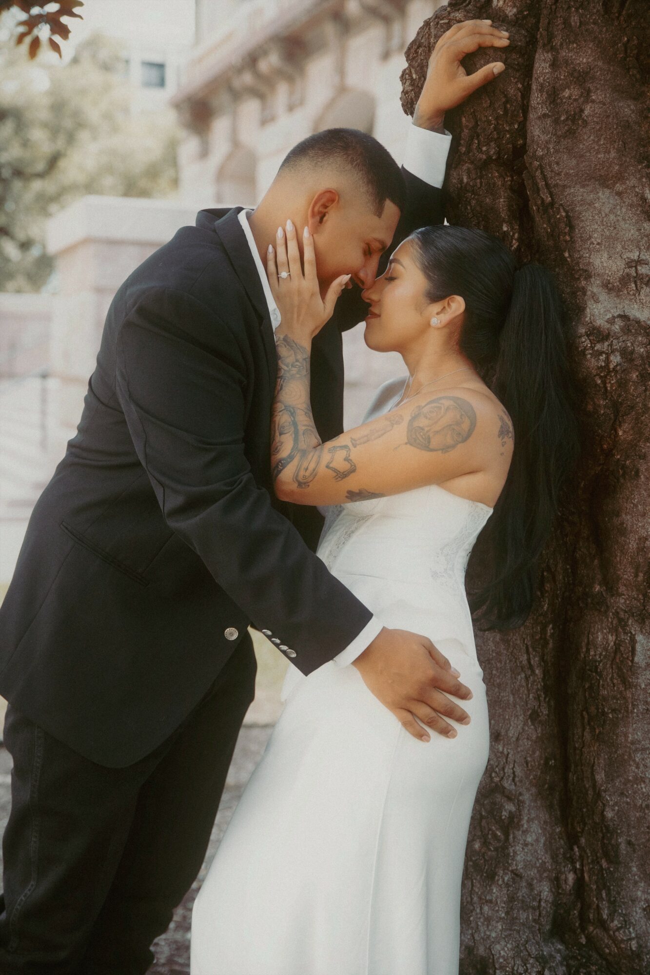 A couple stands close, touching foreheads and holding hands, outdoors near a tree and building, with a romantic atmosphere.