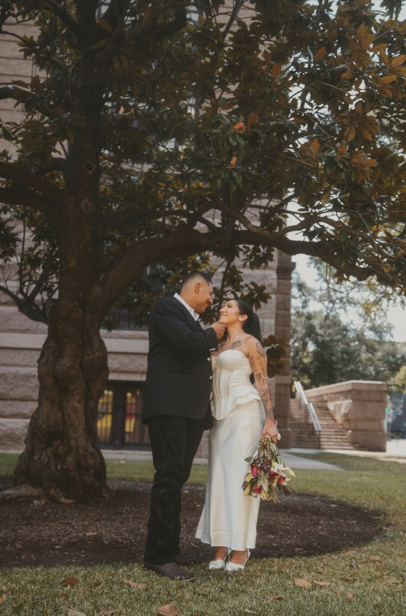 A couple stands under a large tree, with the woman holding a bouquet of flowers, in an outdoor setting.