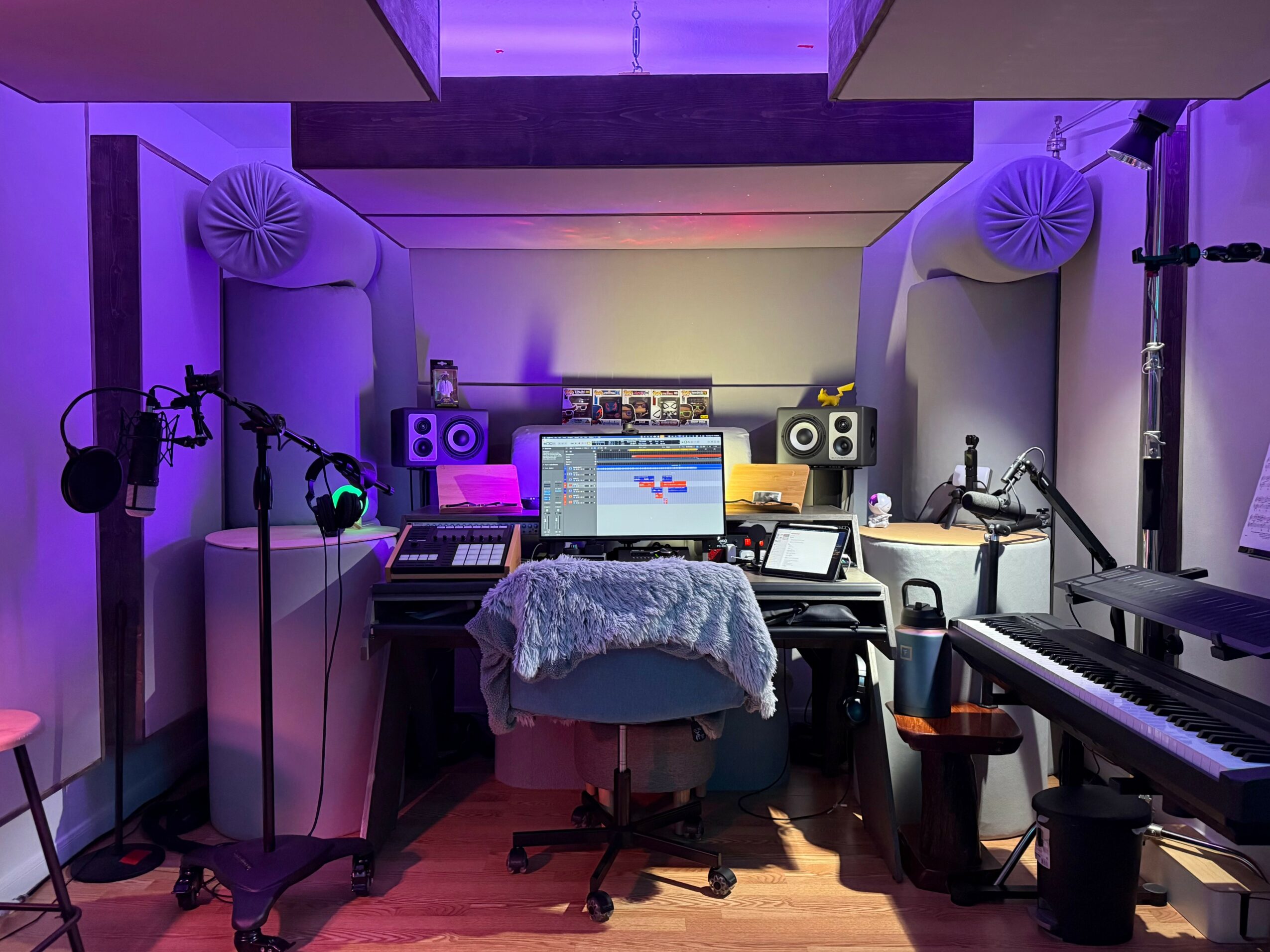 Music studio with computer, speakers, keyboard, and microphones, illuminated by purple and pink lighting, with soundproofing panels.