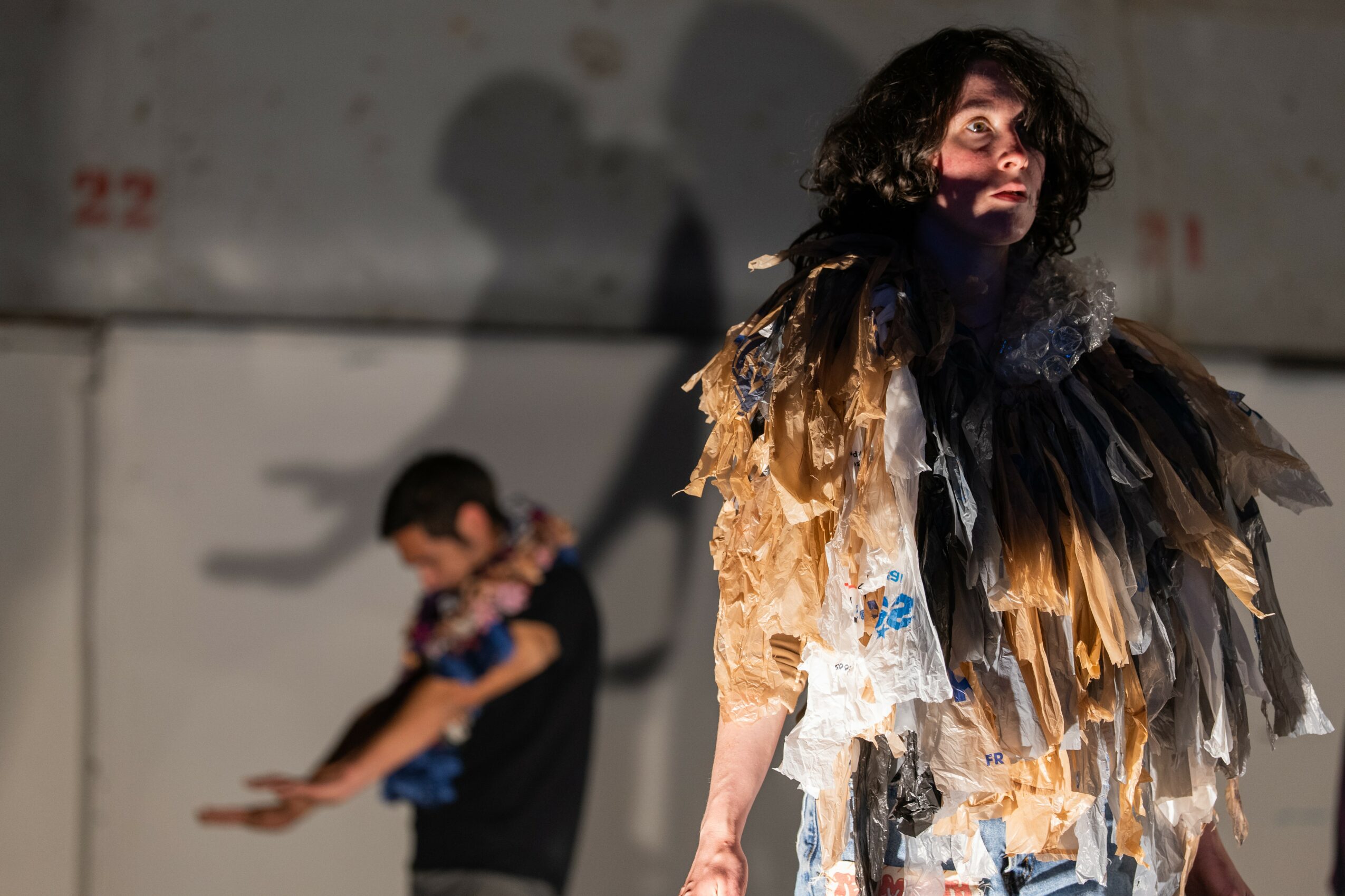 Person with dark curly hair wearing a layered, feathered costume standing indoors with a blurred background.
