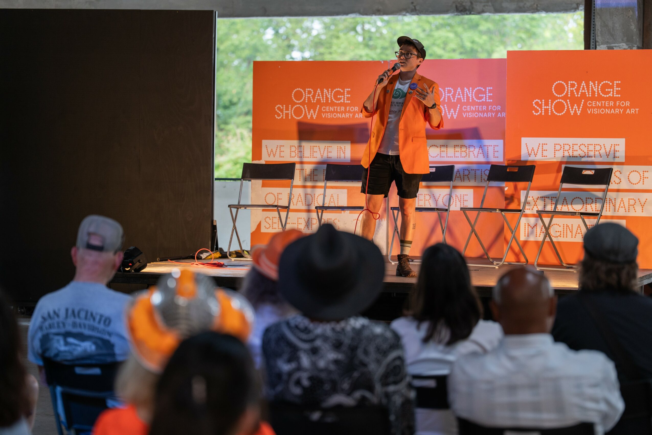 Person on stage speaking into microphone with orange backdrop, audience seated watching, large window behind stage showing trees.