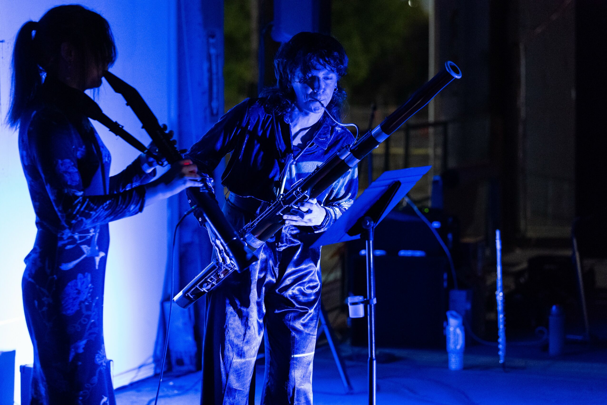 Two musicians perform on stage with blue lighting, one playing a saxophone and the other a bassoon, with music stand nearby.