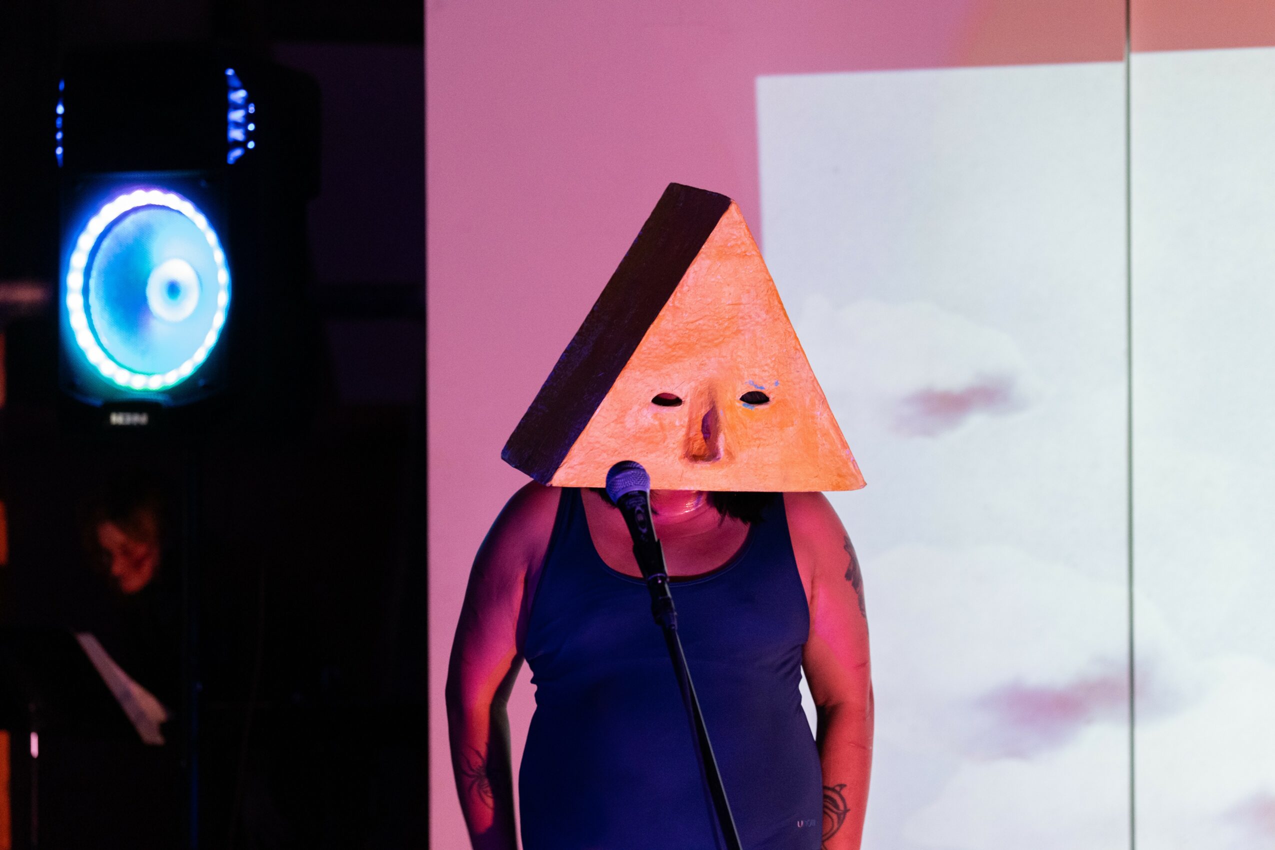 Person wearing a triangular paper hat with a face, standing in front of a pink and white background, speaking into a microphone.
