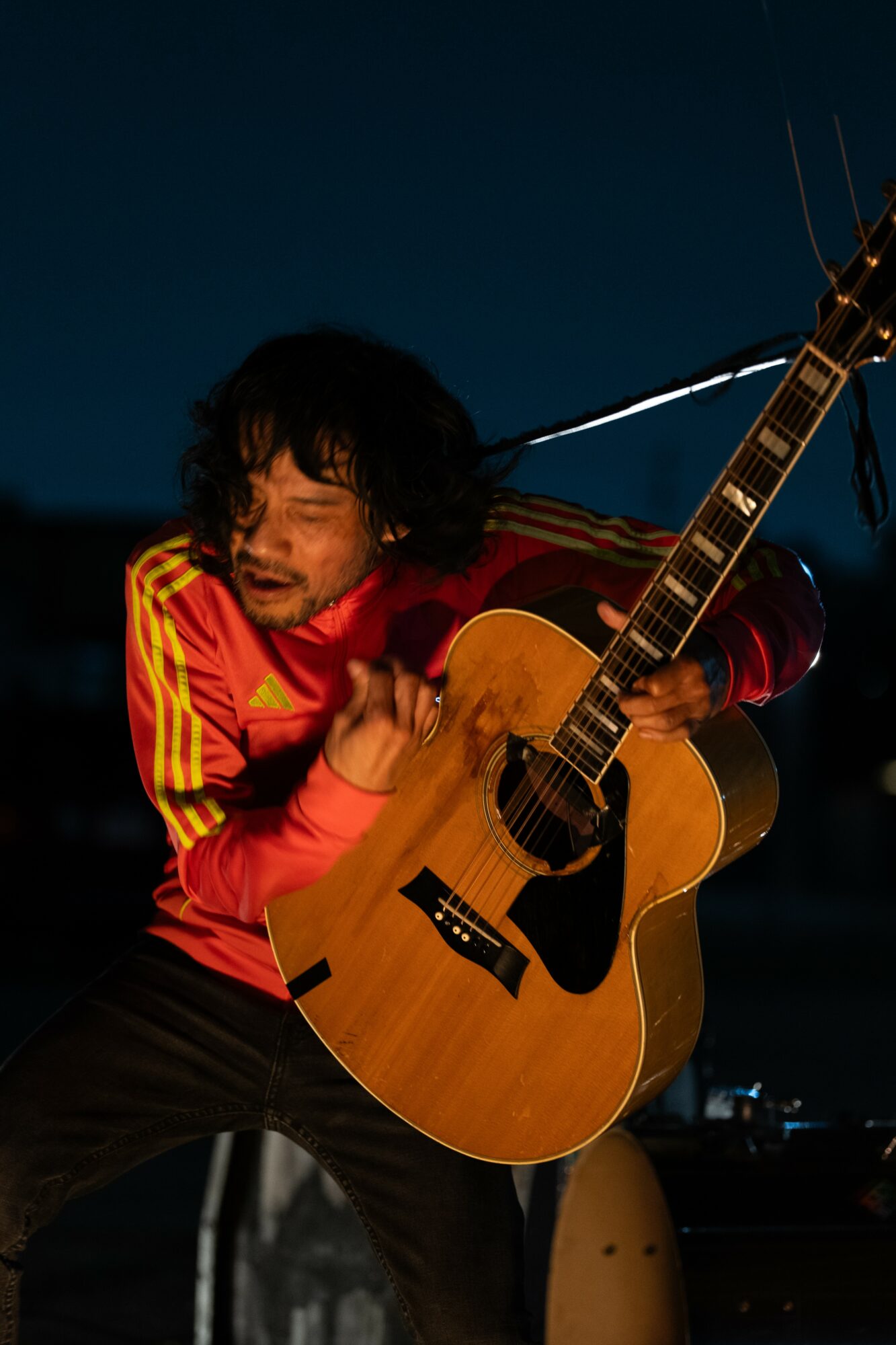 Person with curly hair playing an acoustic guitar outdoors at night, wearing a red and yellow jacket.