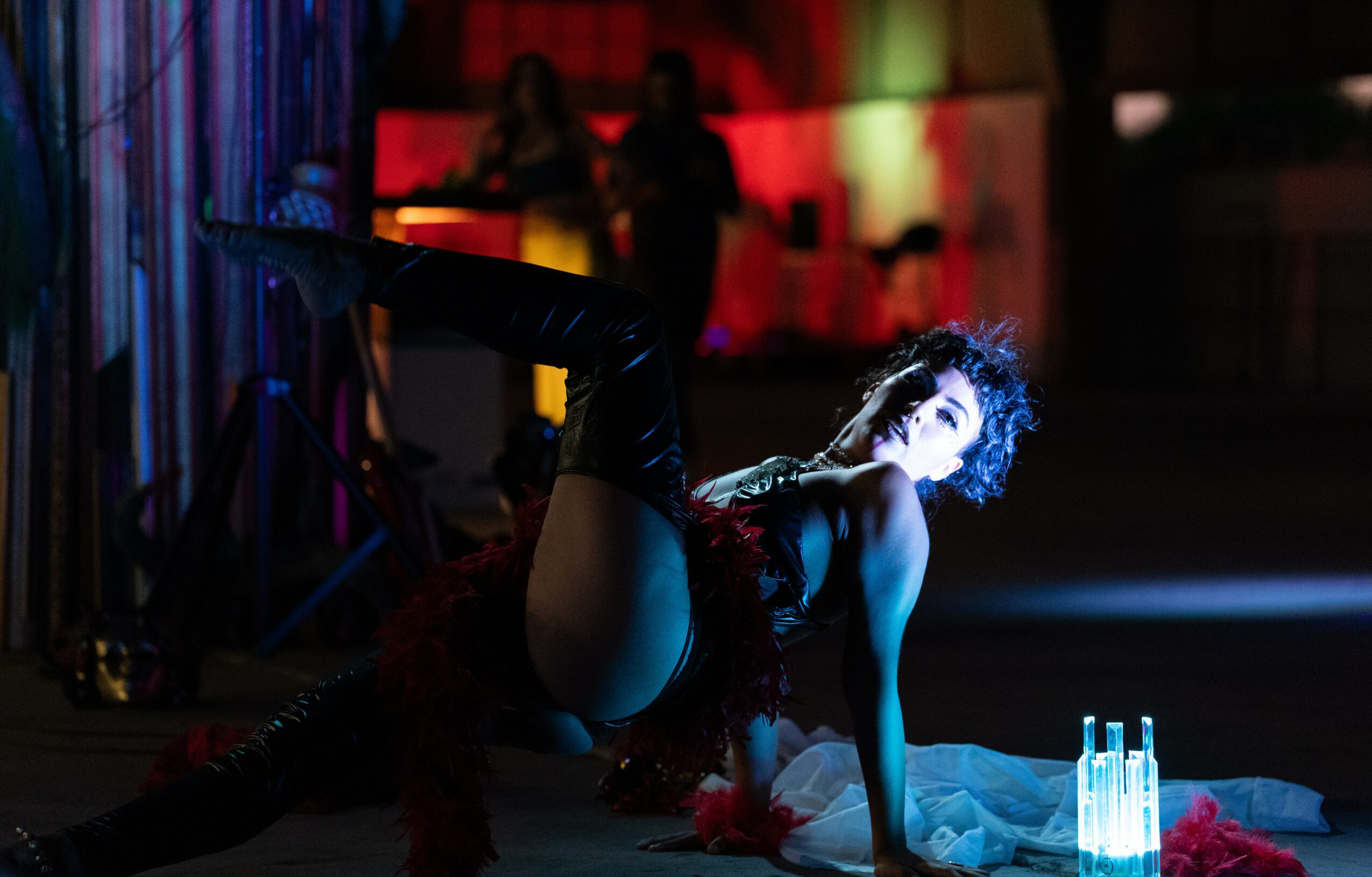 Person in a dark outfit poses on the floor with bottles nearby, illuminated by colorful stage lighting.